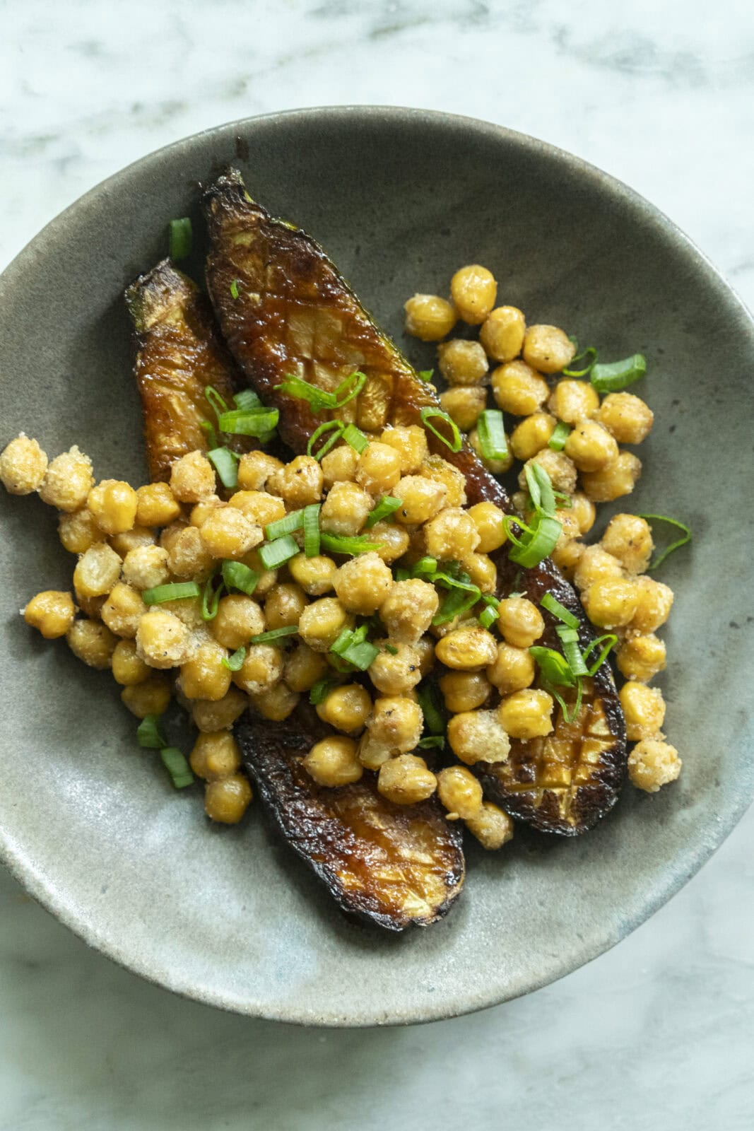 A bowl contains roasted eggplant halves topped with seasoned chickpeas and garnished with chopped green onions.