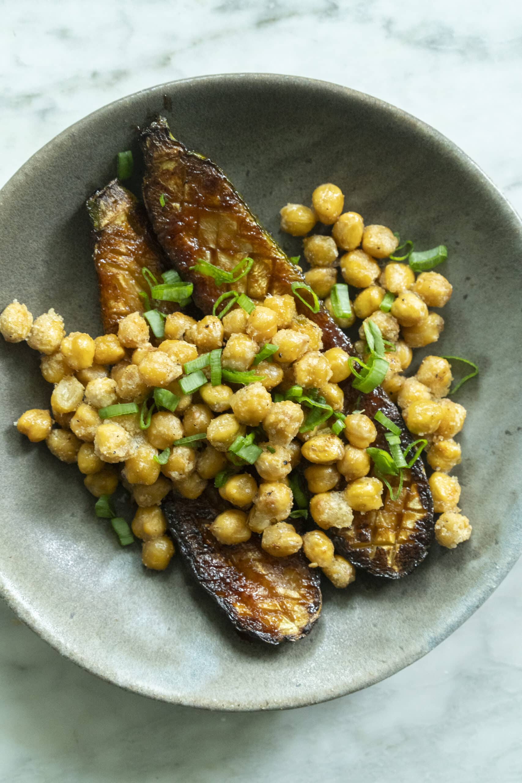 A bowl contains roasted eggplant halves topped with seasoned chickpeas and garnished with chopped green onions.