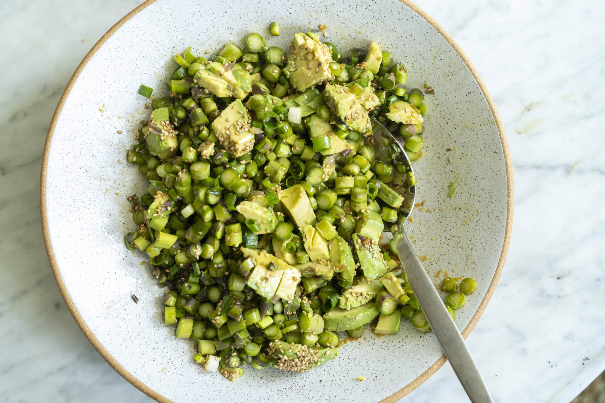 A bowl of chopped asparagus and avocado salad with sesame seeds and a spoon, set on a light marble surface.