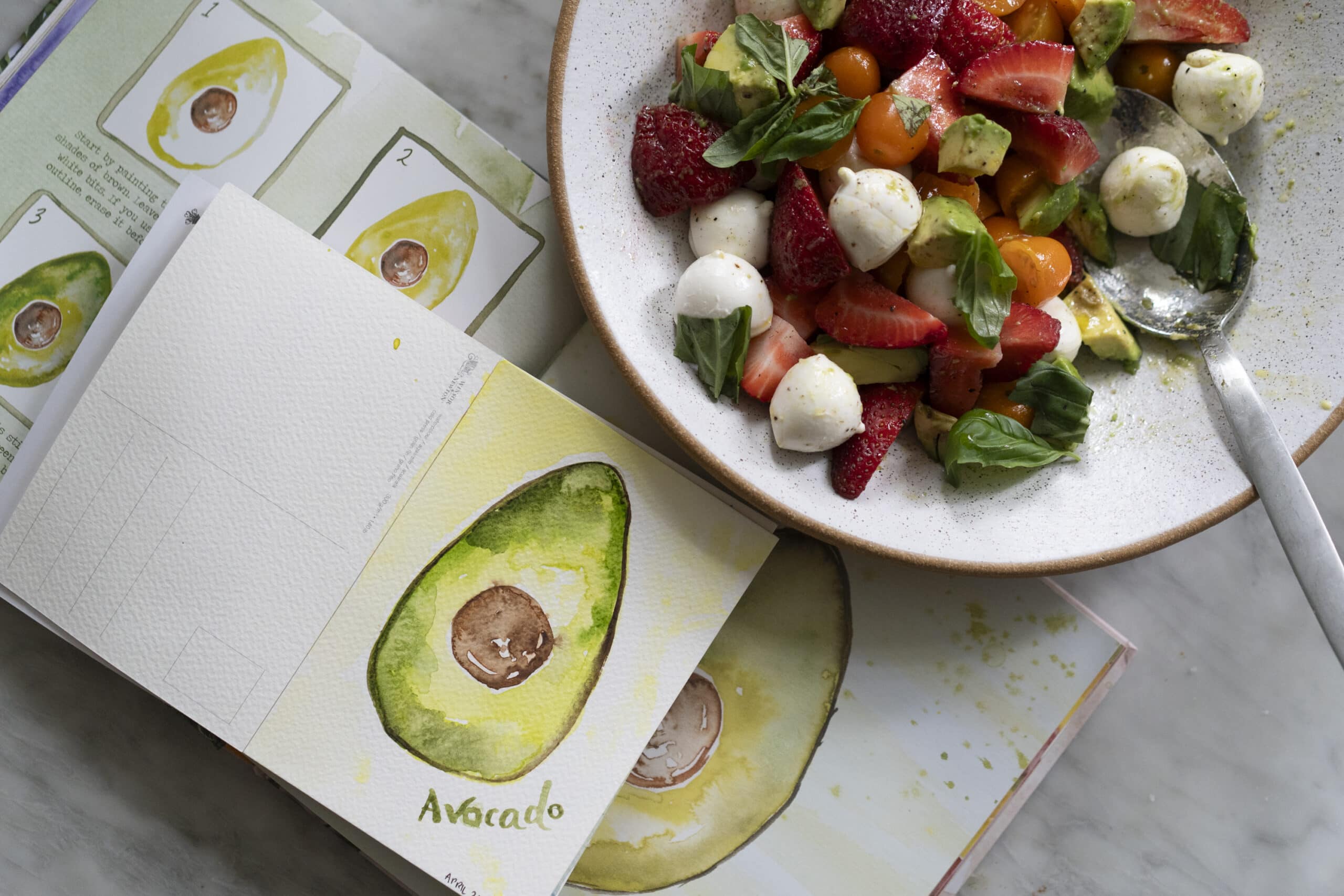 A bowl of salad with strawberries, tomatoes, mozzarella, avocado, and basil next to open sketchbooks featuring avocado illustrations.