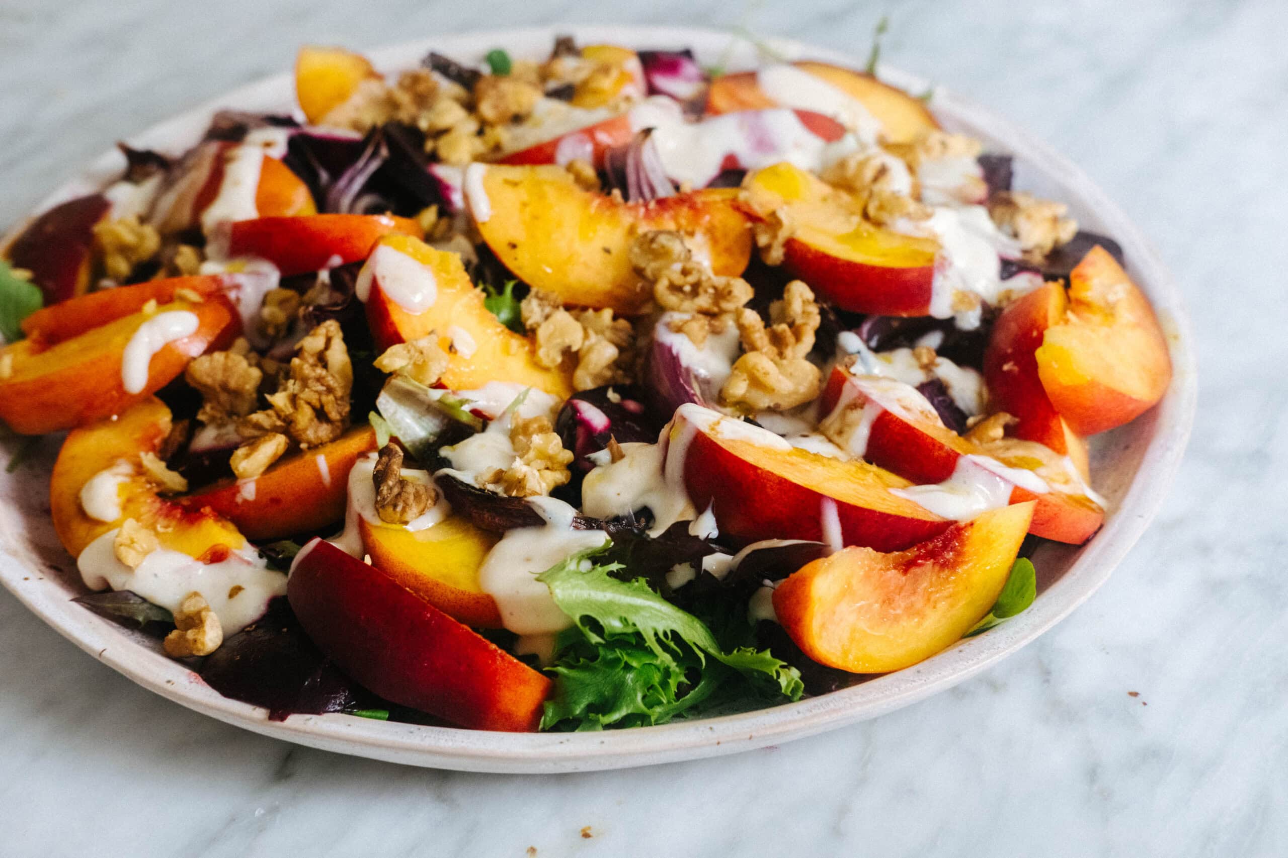 A plate of mixed greens topped with sliced peaches, walnuts, and a drizzle of creamy dressing on a marble surface.