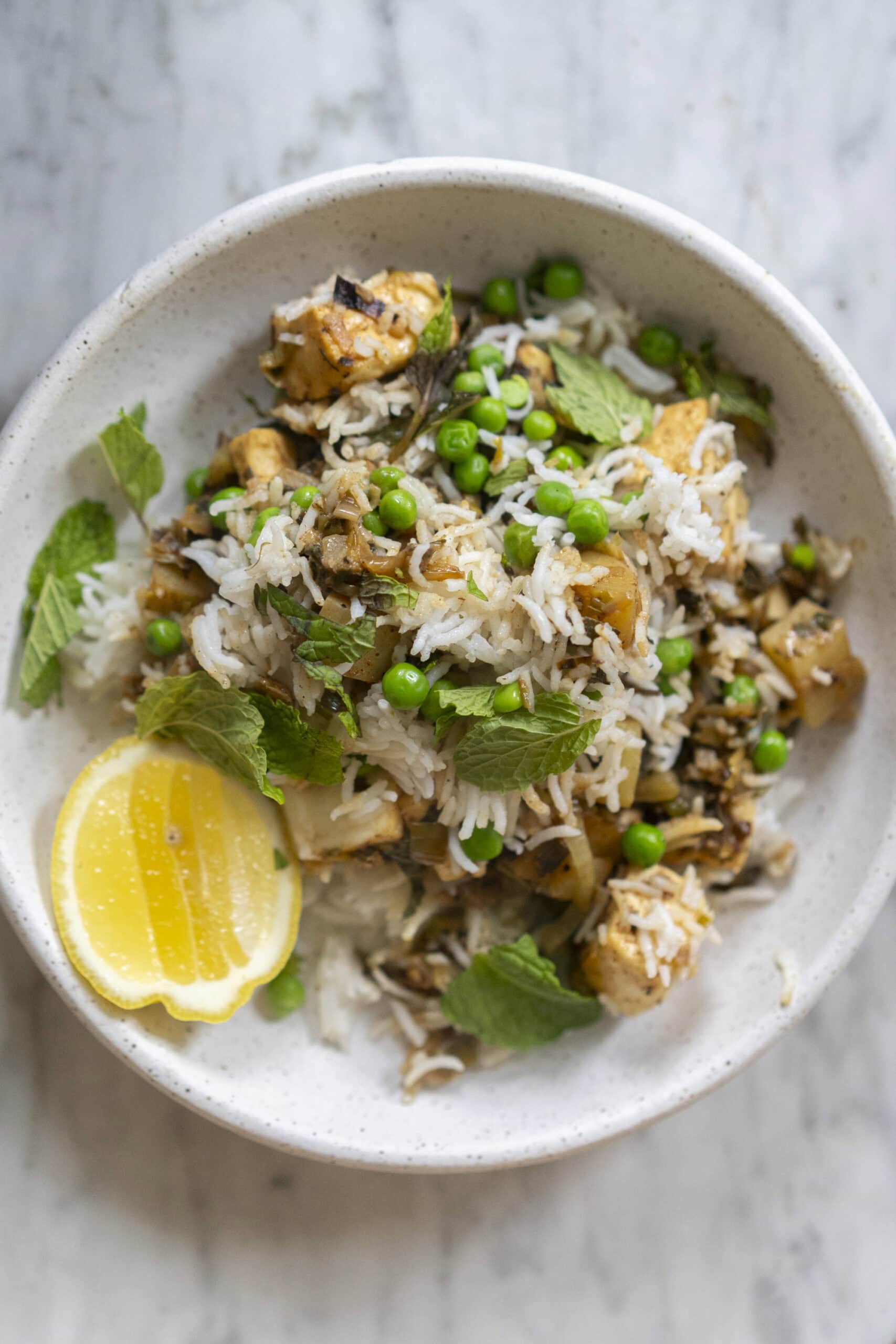 A bowl of rice topped with peas, tofu, mint leaves, and onions, garnished with lemon slices on the side.