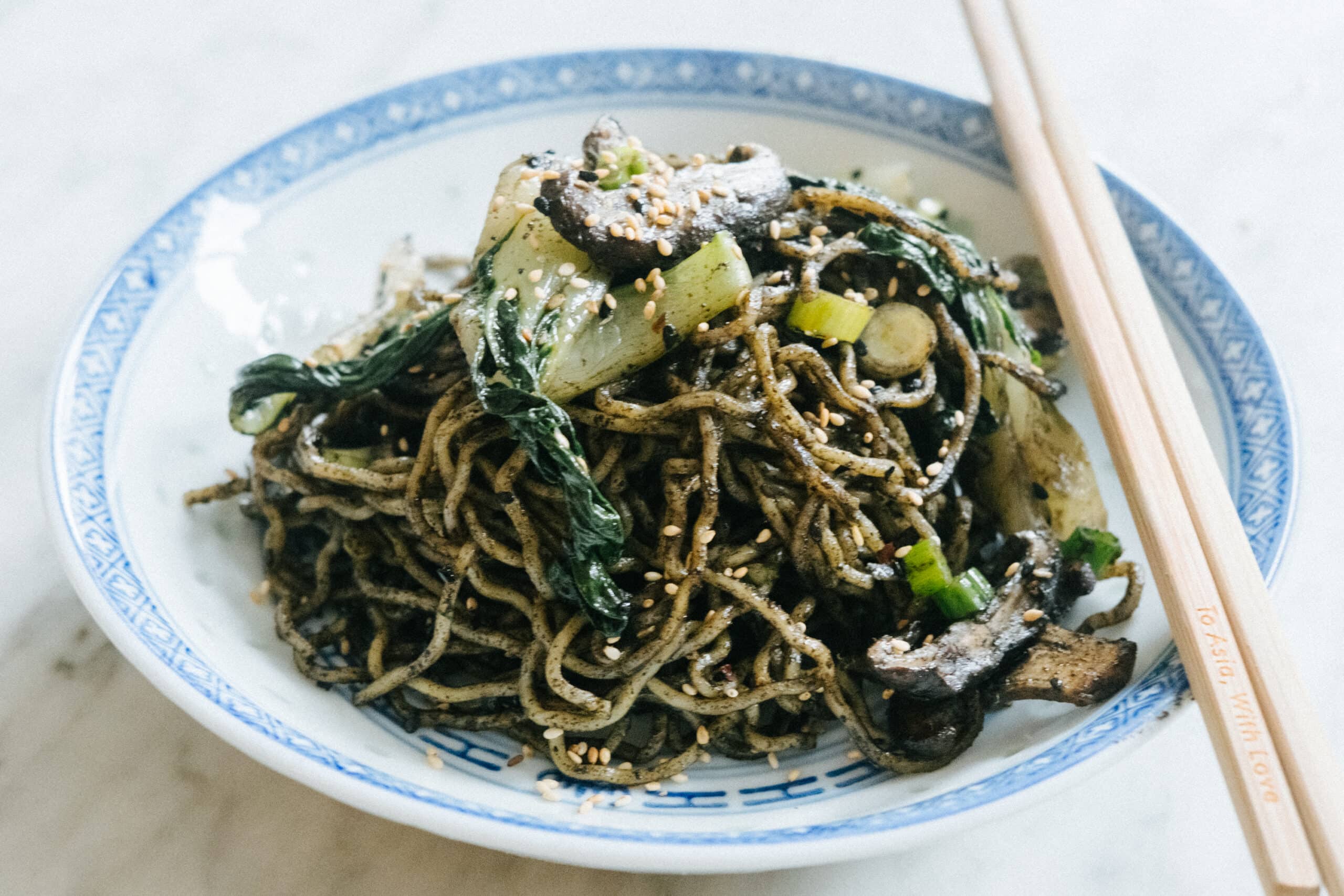 A plate of stir-fried noodles with leafy greens, mushrooms, and sesame seeds, served with chopsticks on a white and blue patterned dish.