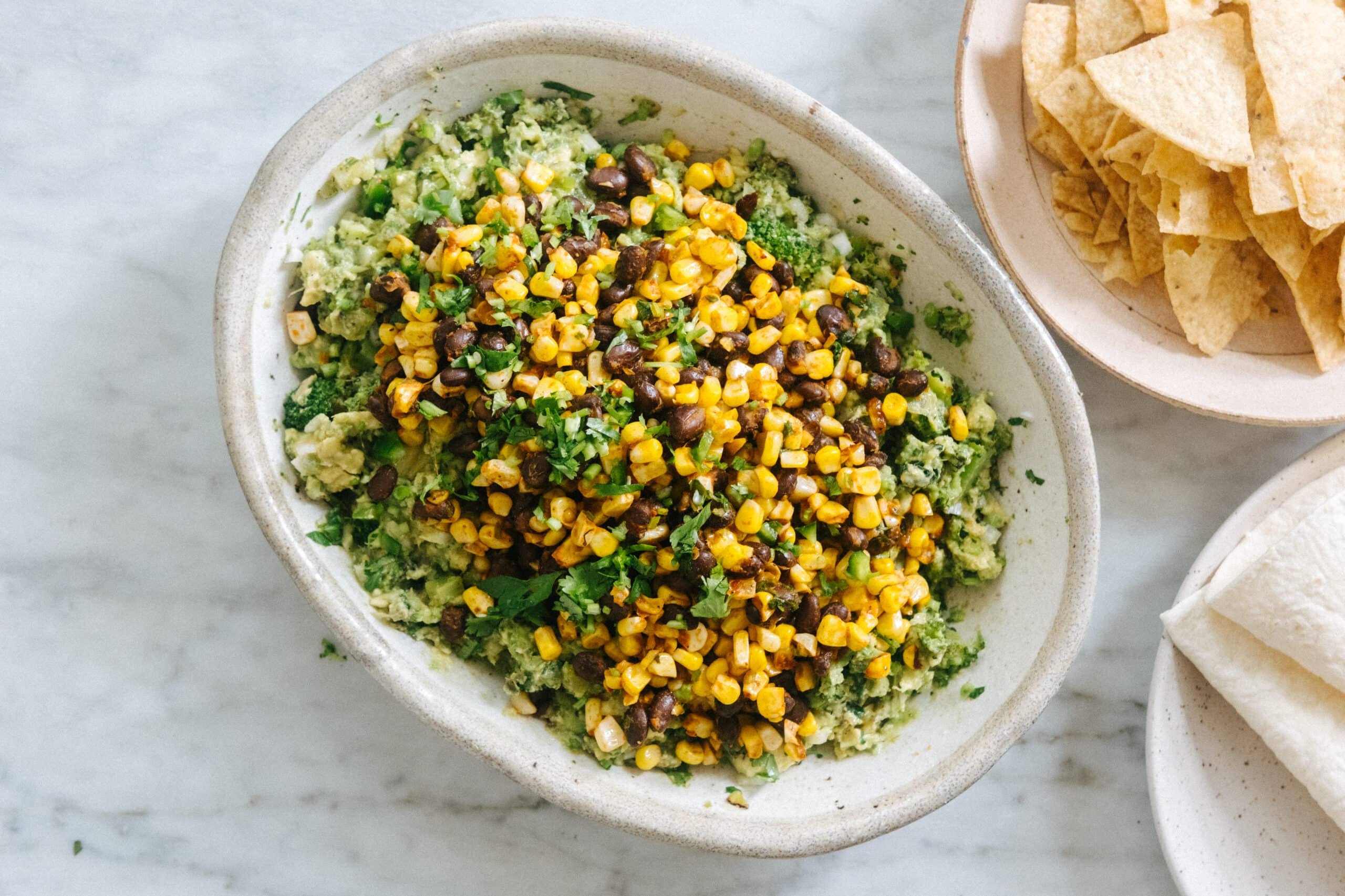 A bowl of guacamole topped with roasted corn, black beans, and chopped cilantro, next to plates of tortilla chips and tortillas on a marble surface.