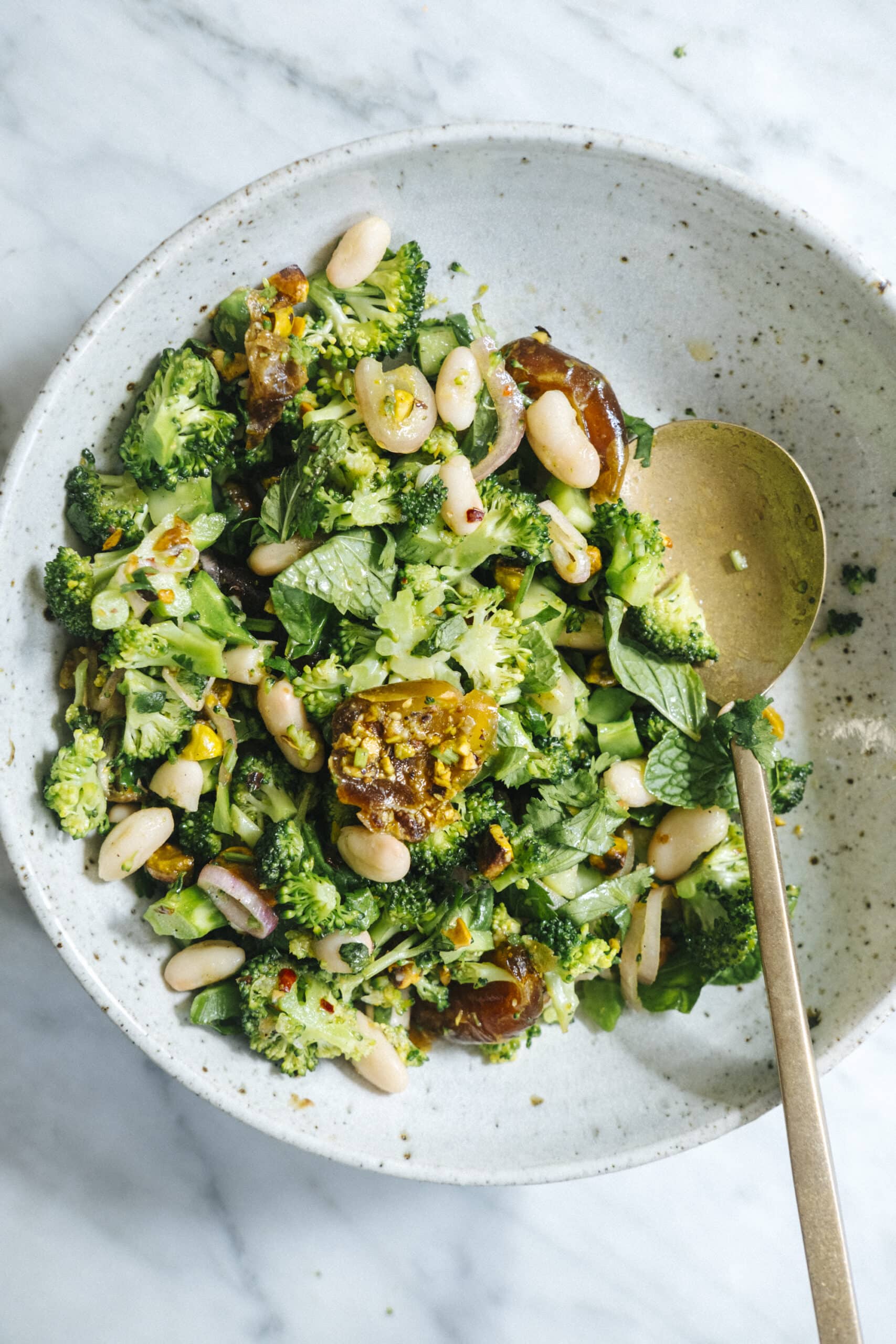A bowl of broccoli salad with white beans, chopped greens, and dates, served with a gold spoon on a white plate.