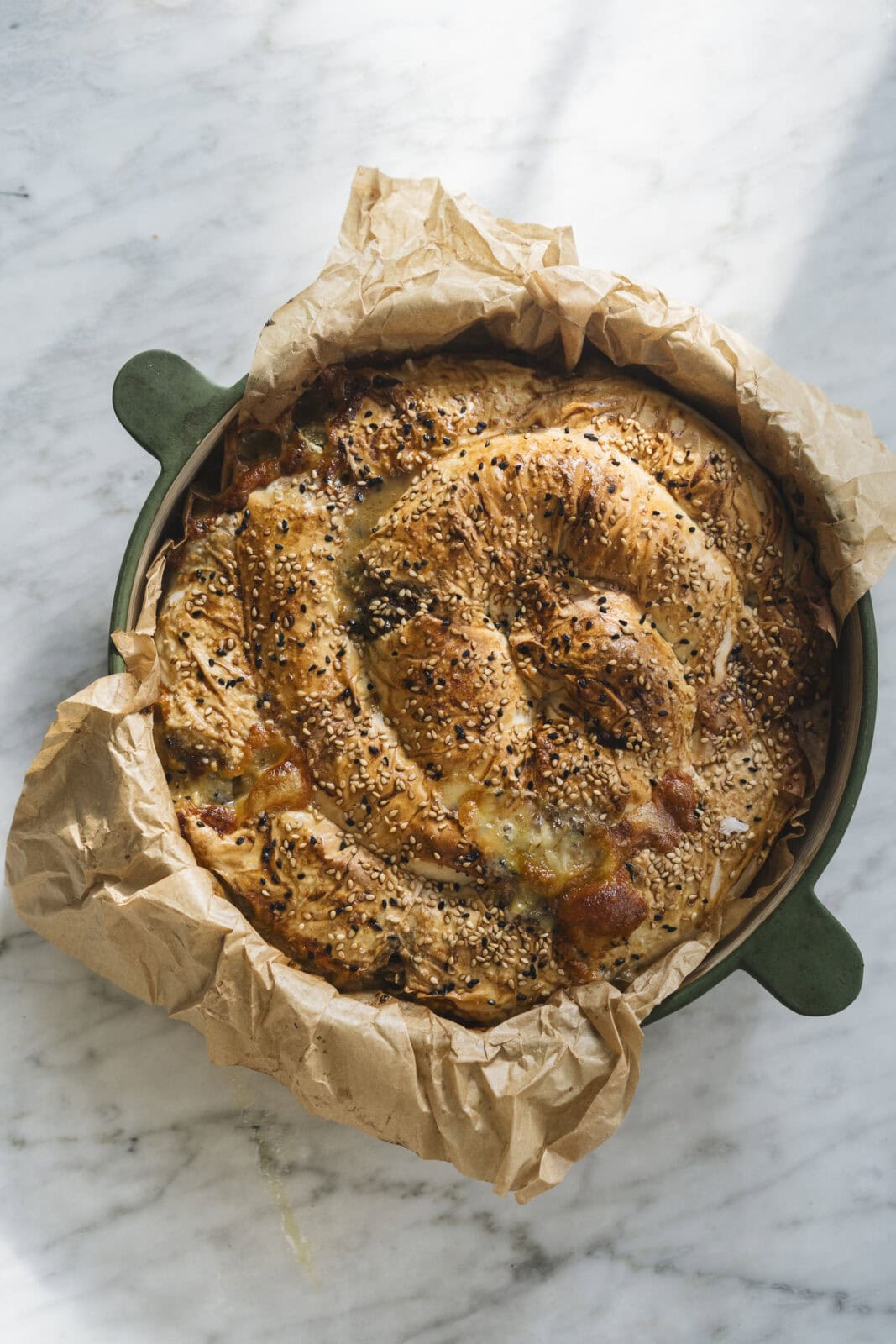 A round, golden-brown savory pastry topped with sesame and black seeds, baked in a green dish lined with crumpled parchment paper on a marble surface.