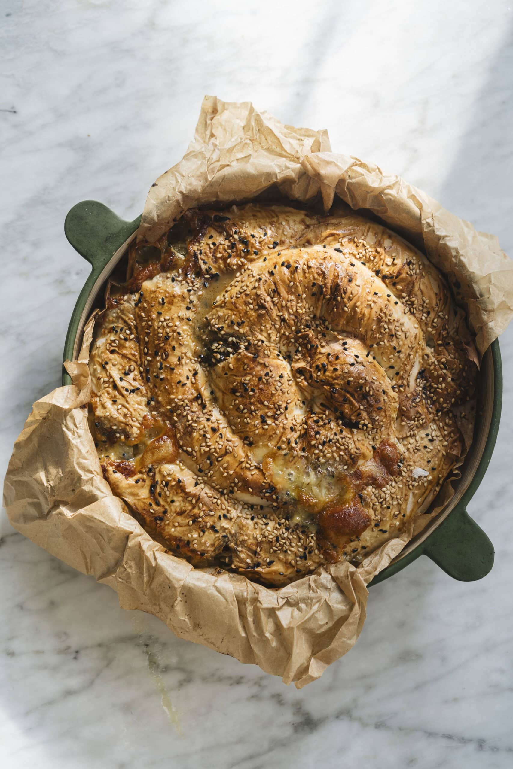 A round, golden-brown savory pastry topped with sesame and black seeds, baked in a green dish lined with crumpled parchment paper on a marble surface.