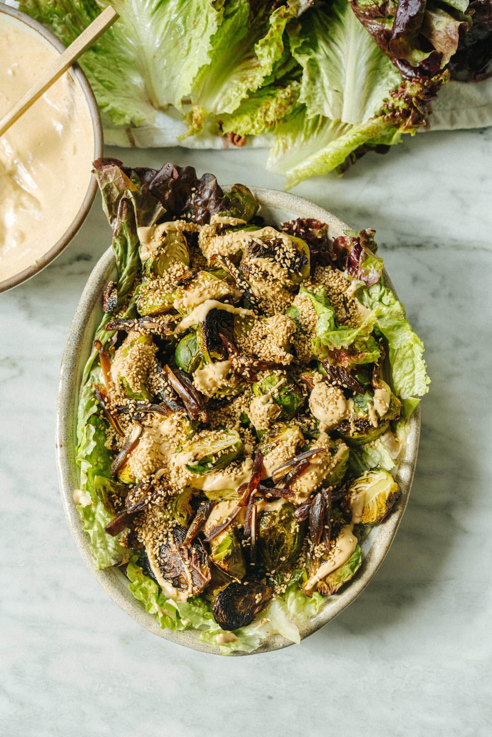 A bowl of roasted Brussels sprouts and lettuce topped with creamy dressing and sesame seeds, with a side of dressing and loose lettuce leaves in the background.
