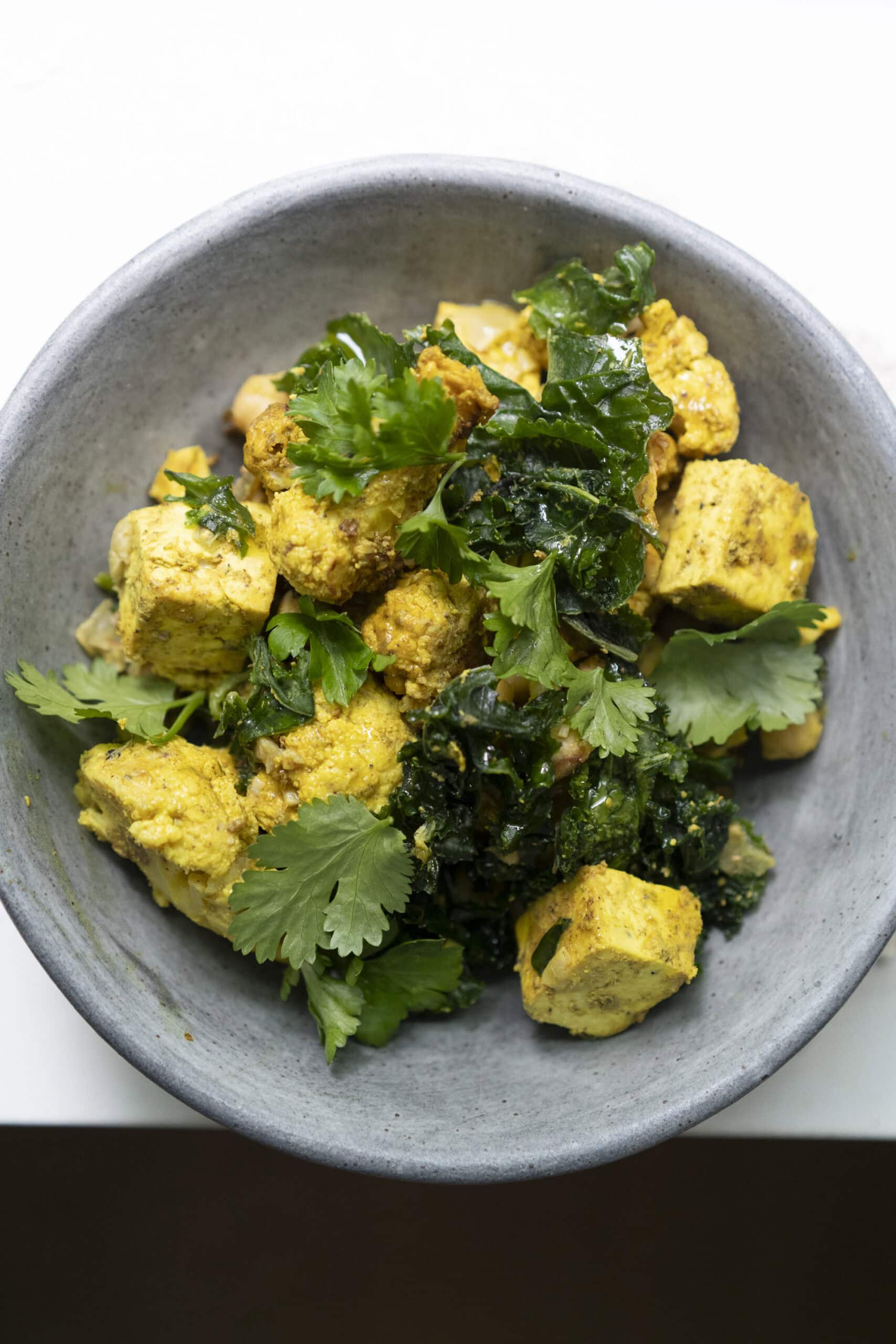 A gray bowl containing yellow tofu cubes, cauliflower pieces, sautéed greens, and topped with fresh cilantro leaves.