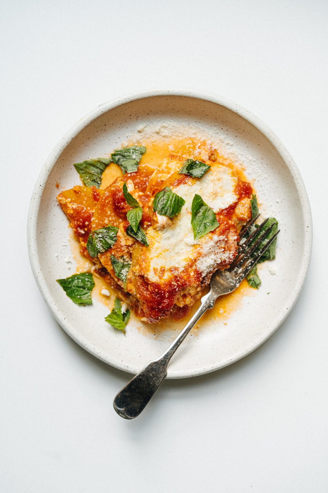 A serving of lasagna topped with tomato sauce, melted cheese, and fresh basil in a ceramic bowl with a fork. - Hetty Lui McKinnon