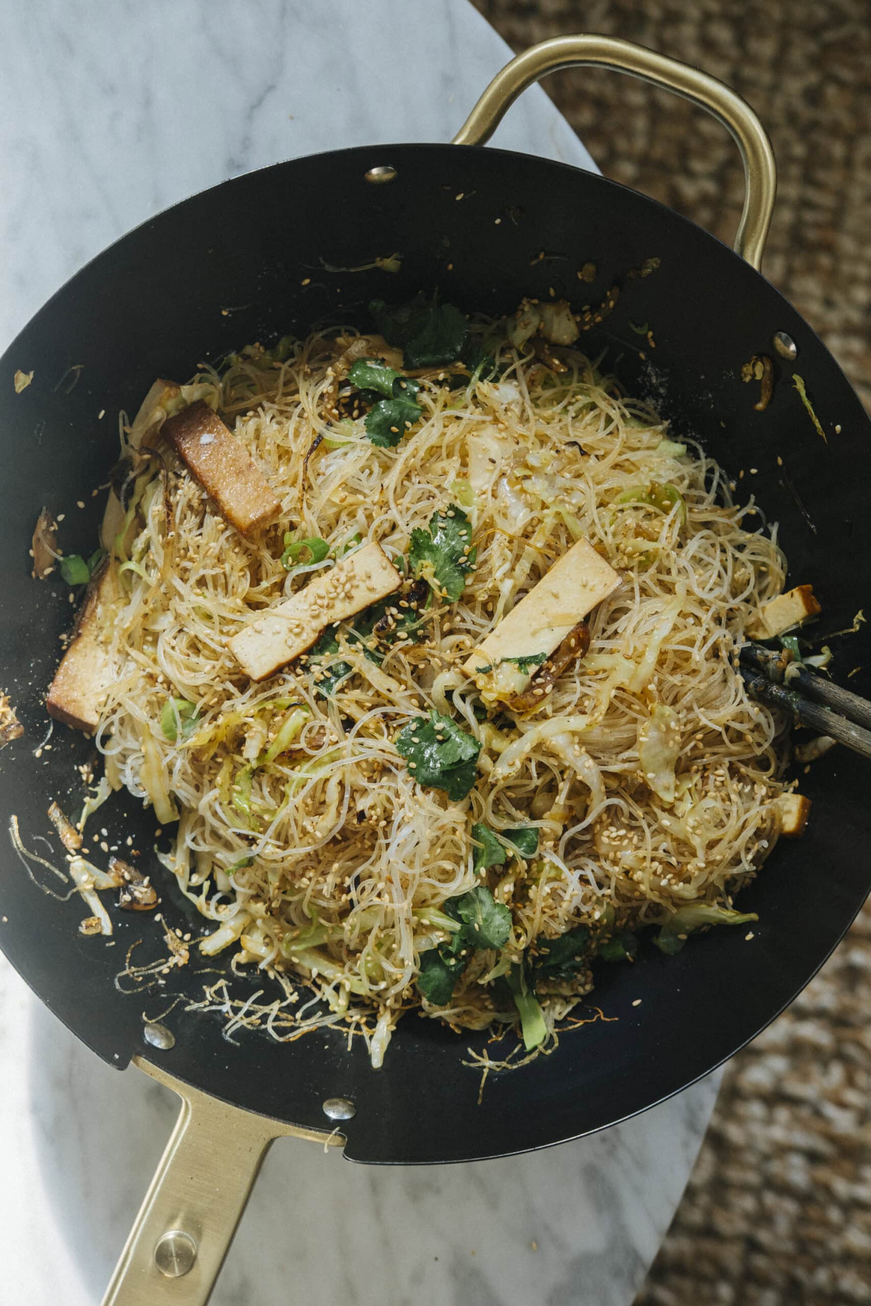 A wok filled with stir-fried rice vermicelli noodles, tofu strips, leafy greens, and sesame seeds, placed on a marble surface.