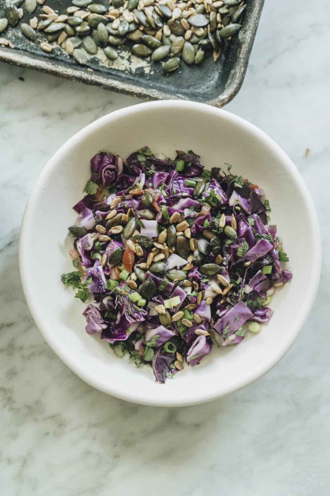 A white bowl filled with chopped red cabbage salad topped with seeds sits on a marble surface, with a tray of more seeds visible in the background.
