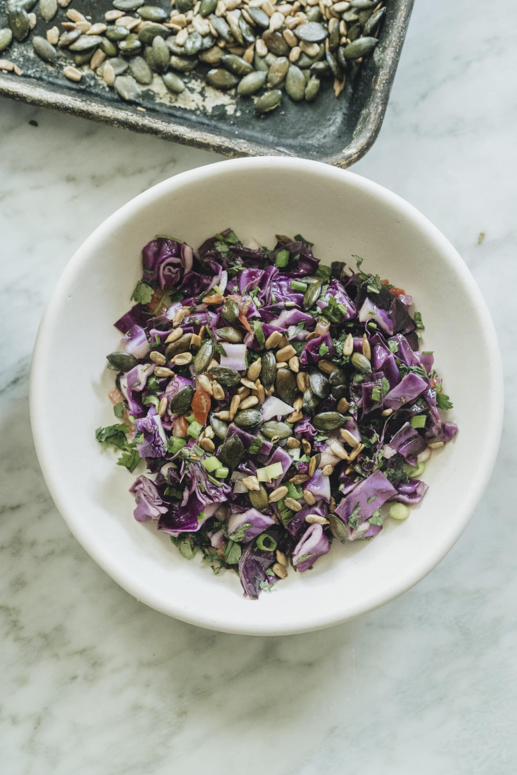 A white bowl filled with chopped red cabbage salad topped with seeds sits on a marble surface, with a tray of more seeds visible in the background.