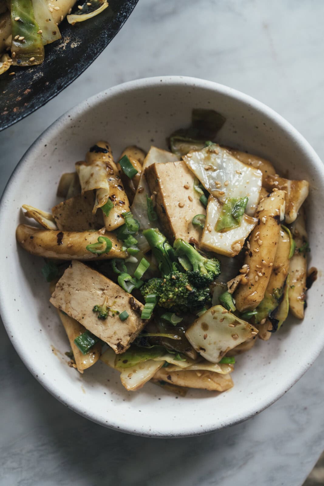 A bowl of stir-fried tofu, rice cakes, broccoli, cabbage, and scallions, garnished with sesame seeds.
