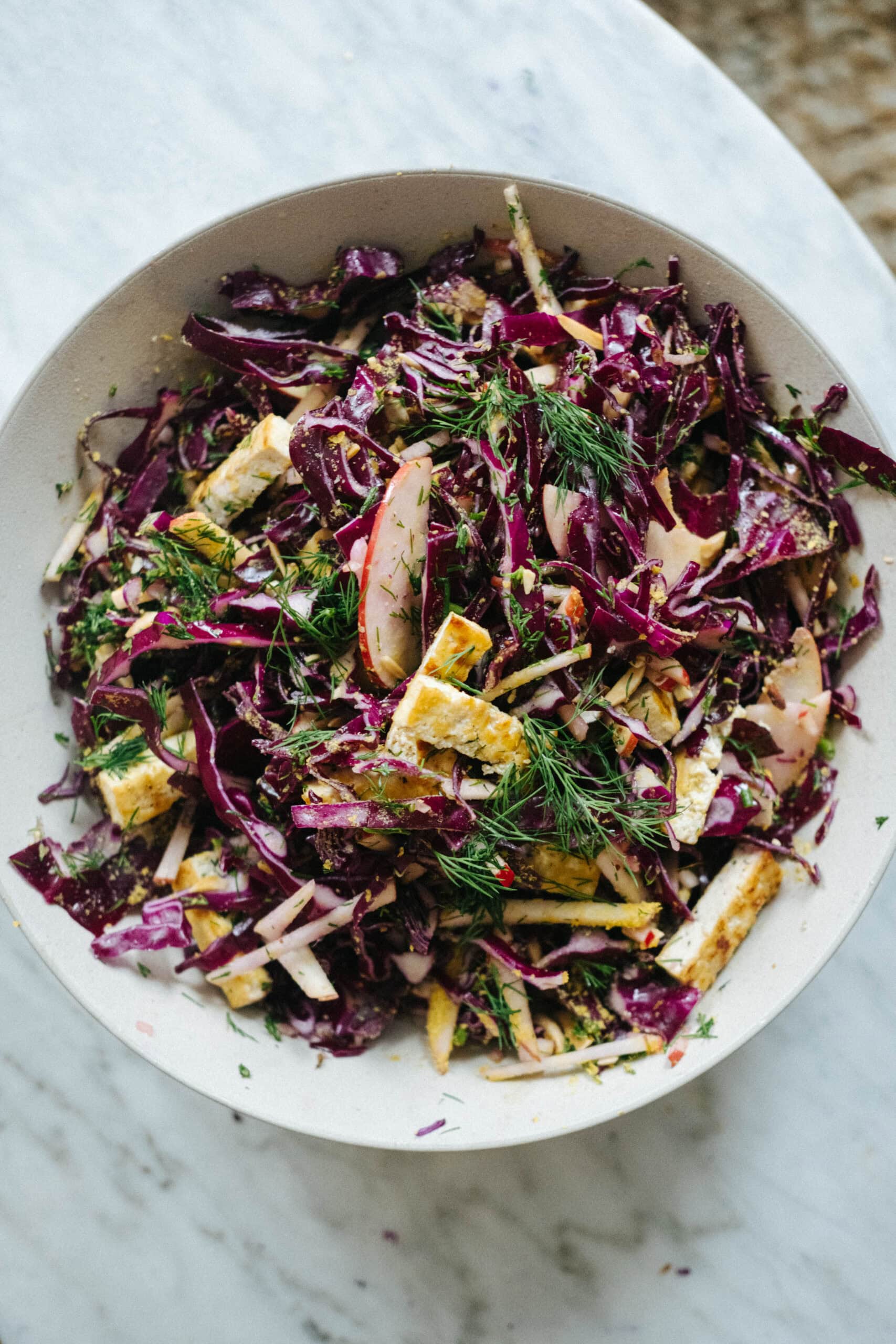 A bowl of red cabbage slaw with grilled tofu slices, fresh dill, and thinly sliced apples on a marble surface.