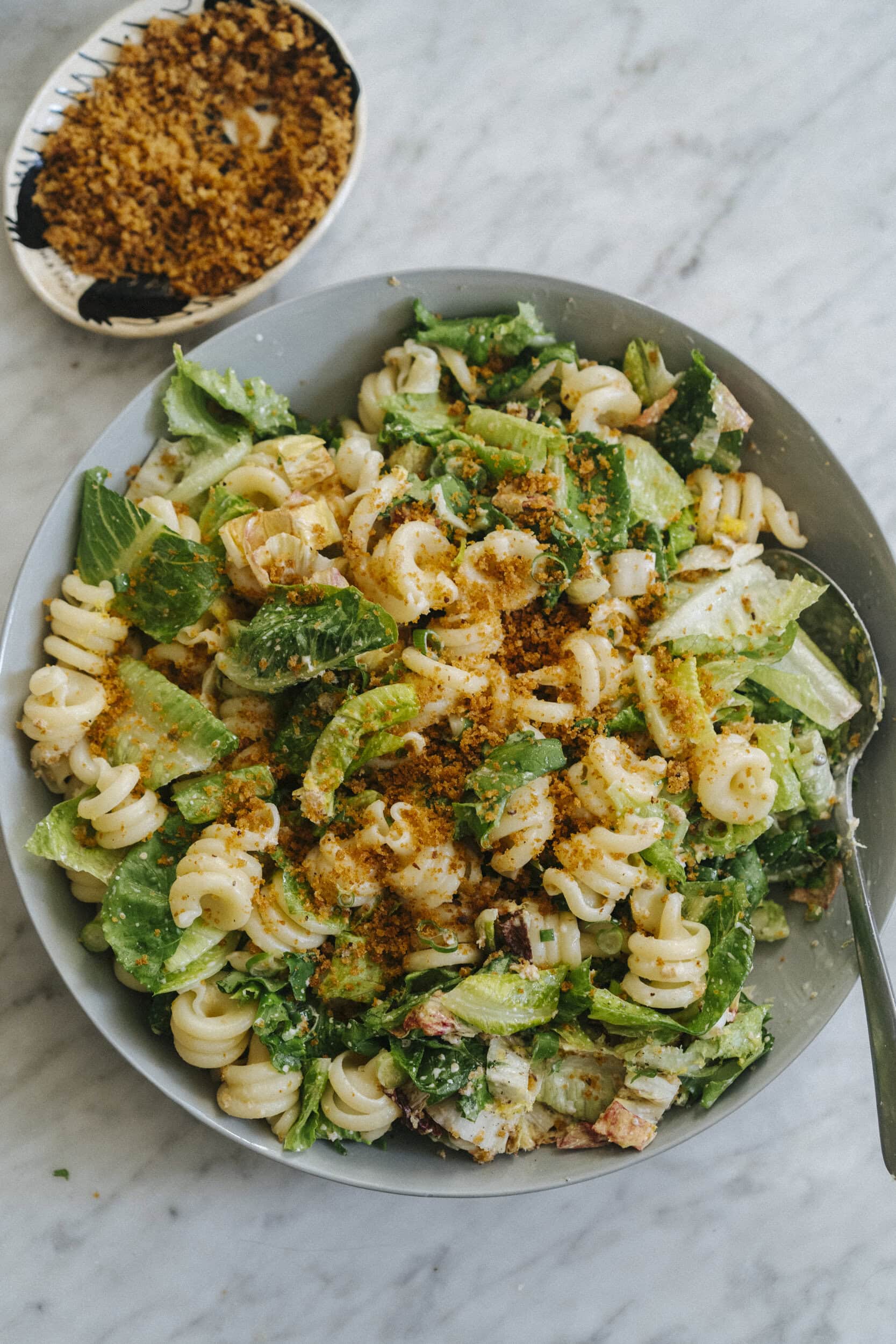 A bowl of pasta salad with lettuce, creamy dressing, and breadcrumbs on top, placed on a marble surface with extra breadcrumbs in a small dish nearby.
