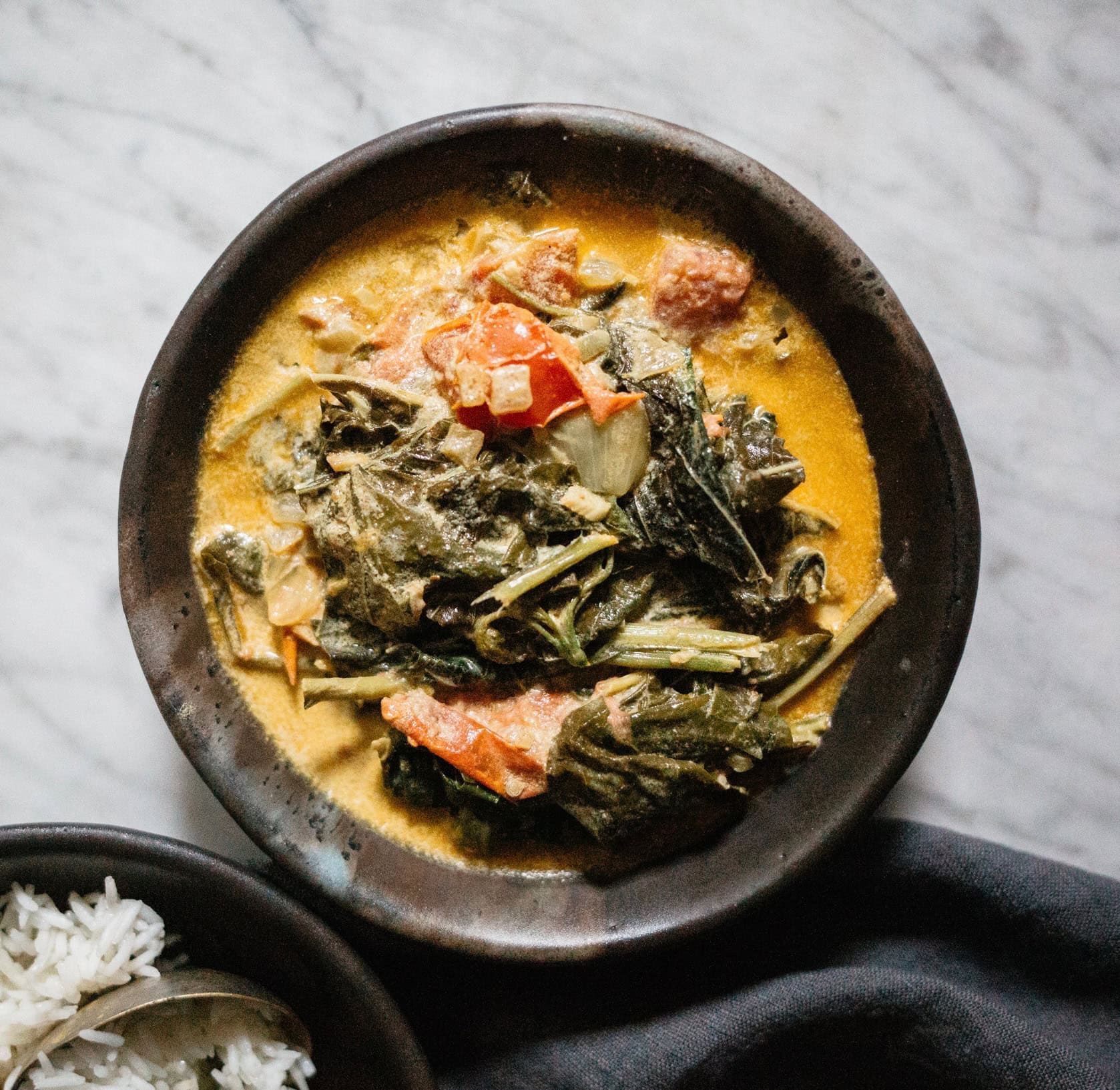 A bowl of vegetable curry in a creamy yellow sauce, with leafy greens and diced vegetables, sits next to a bowl of white rice on a marble surface. - Hetty Lui McKinnon
