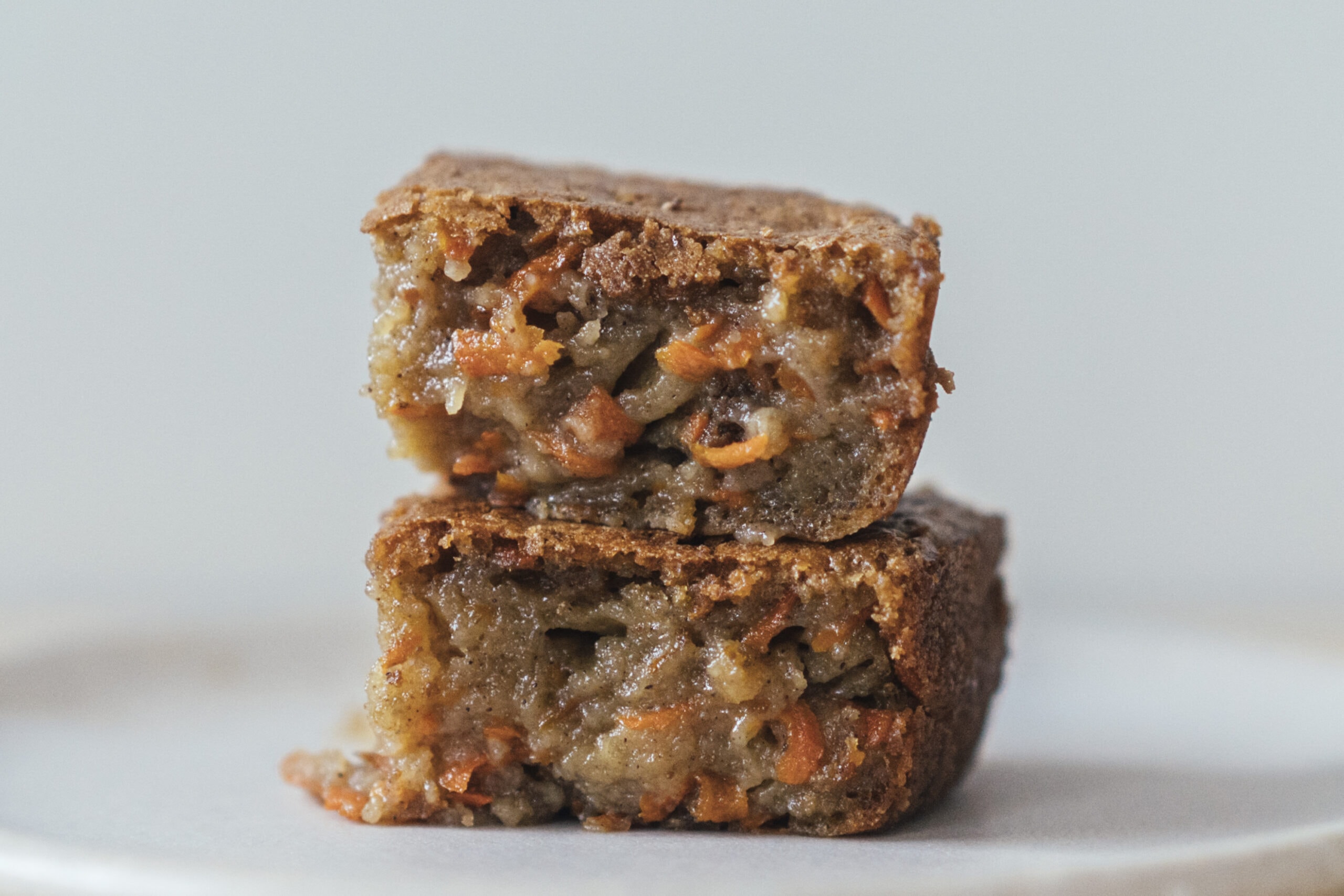 Two pieces of moist carrot cake bars stacked on a white plate, showing a dense, textured interior with visible carrot pieces.