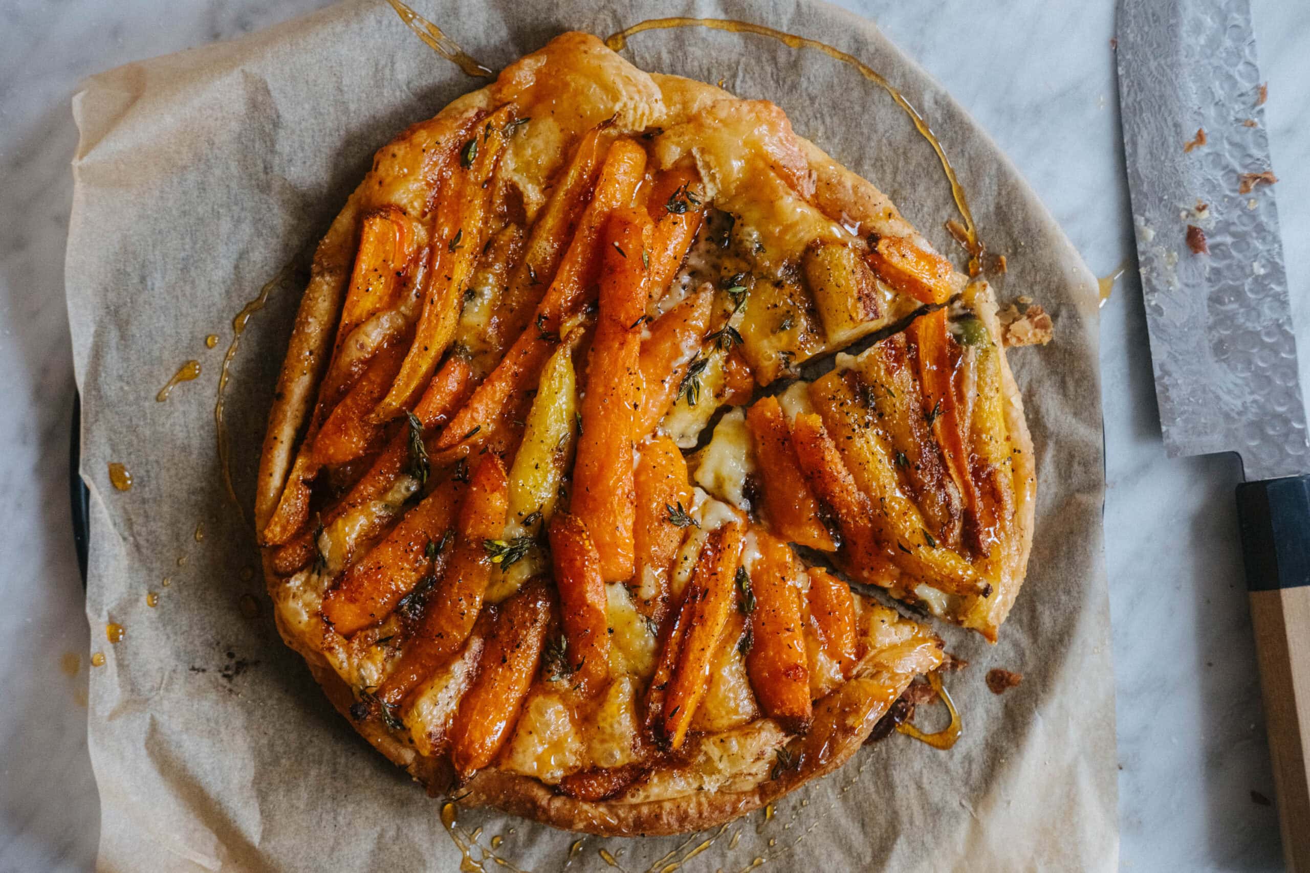A rustic carrot tart with golden pastry, sliced and arranged on parchment paper, sits on a marble surface next to a knife.
