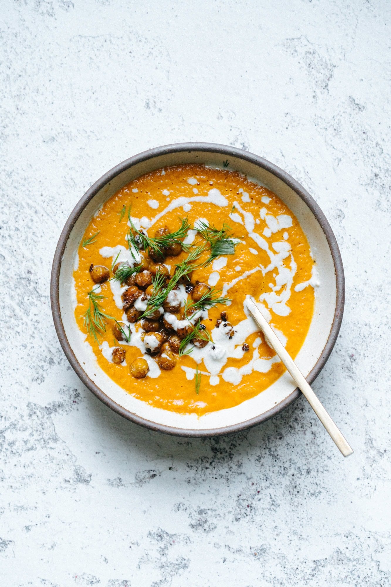 Bowl of orange soup garnished with roasted chickpeas, fresh herbs, and a drizzle of cream, with a spoon resting inside.