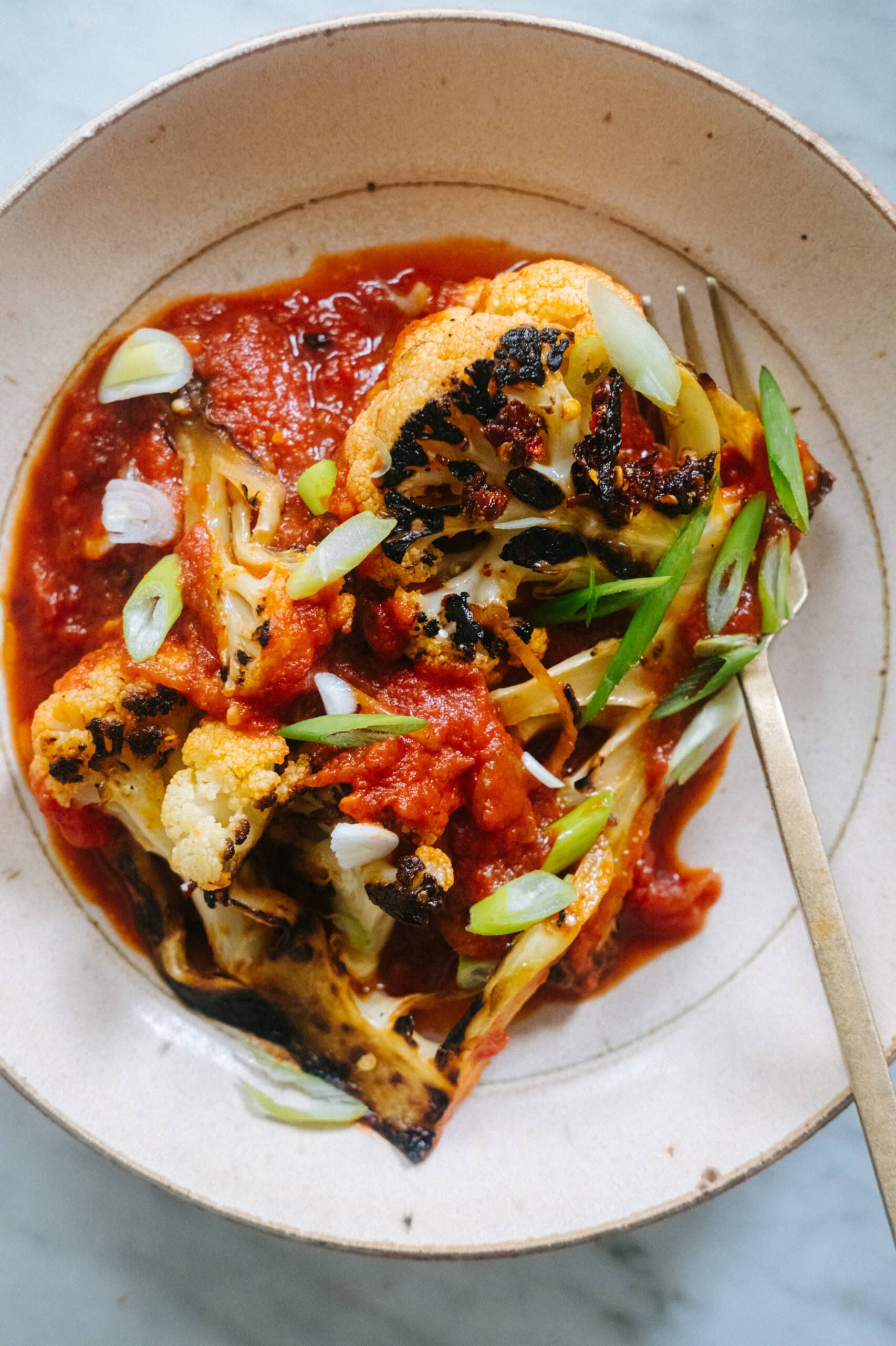 A plate with roasted cauliflower steaks topped with tomato sauce and sliced green onions, served with a fork.