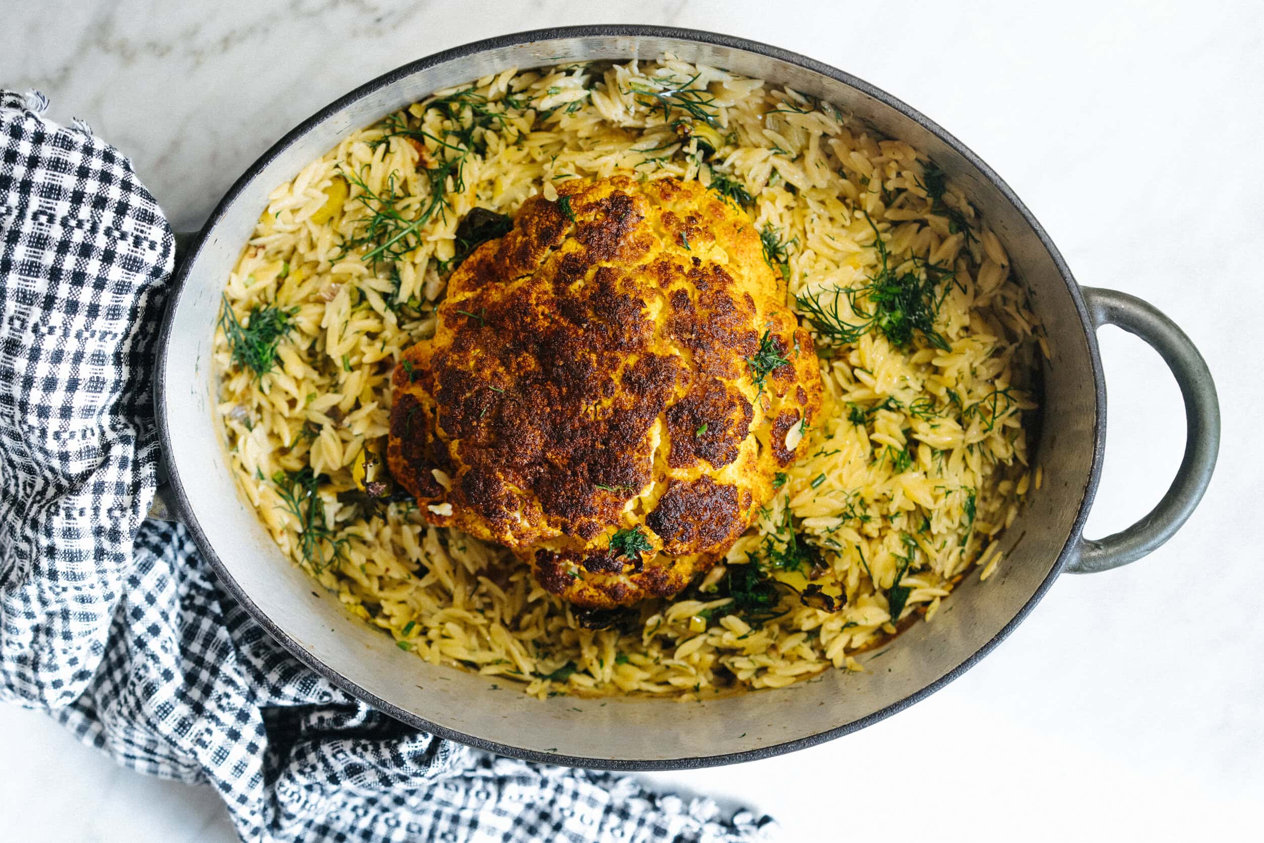 A whole roasted cauliflower sits on a bed of orzo pasta with herbs in an oval baking dish on a marble surface next to a checkered towel.