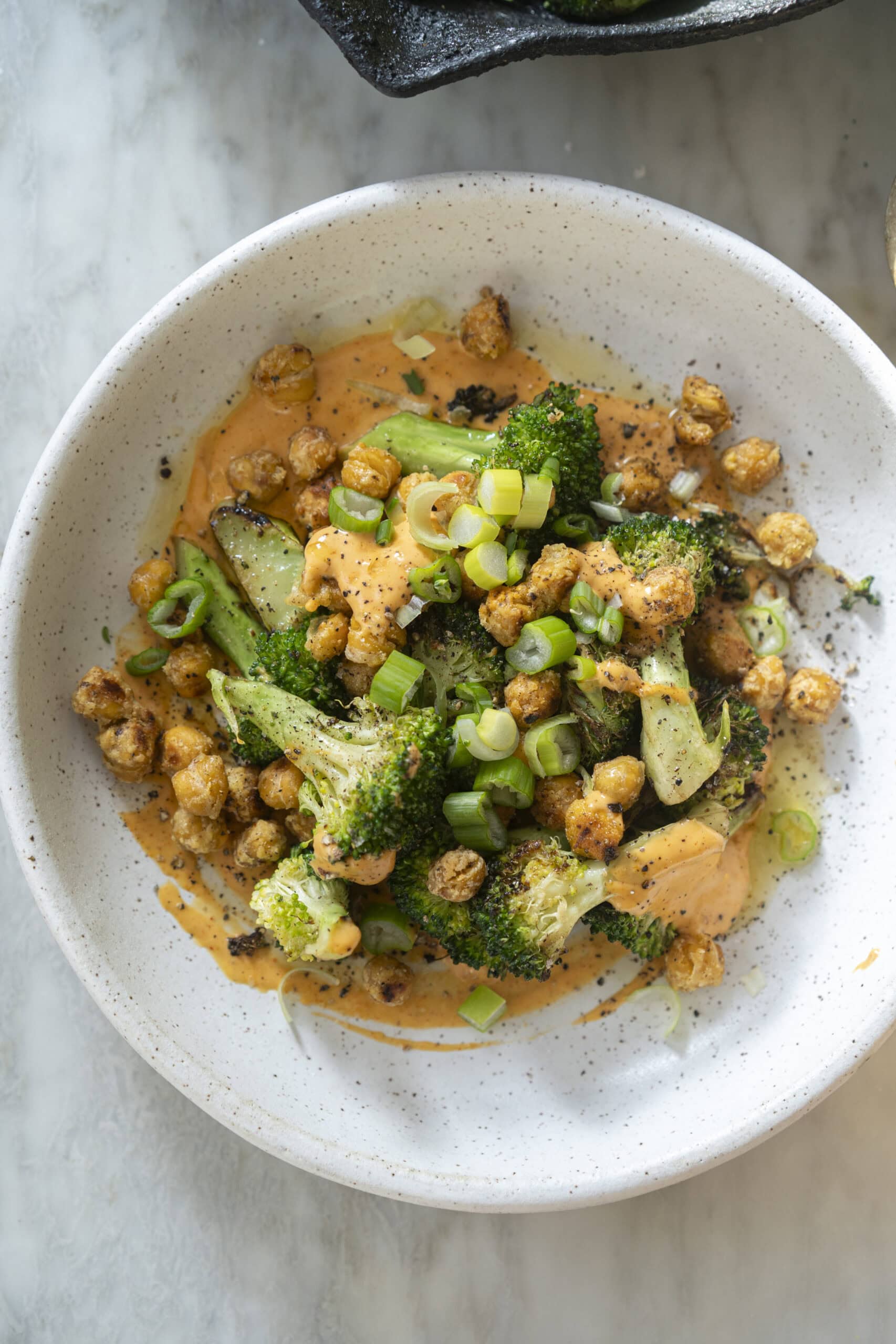 A white bowl filled with roasted broccoli, chickpeas, sliced green onions, and an orange-colored sauce on a marble surface.