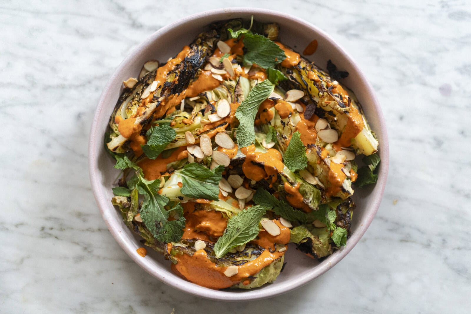A bowl of grilled lettuce topped with sliced almonds, fresh mint leaves, and drizzled with an orange-colored sauce on a marble surface.