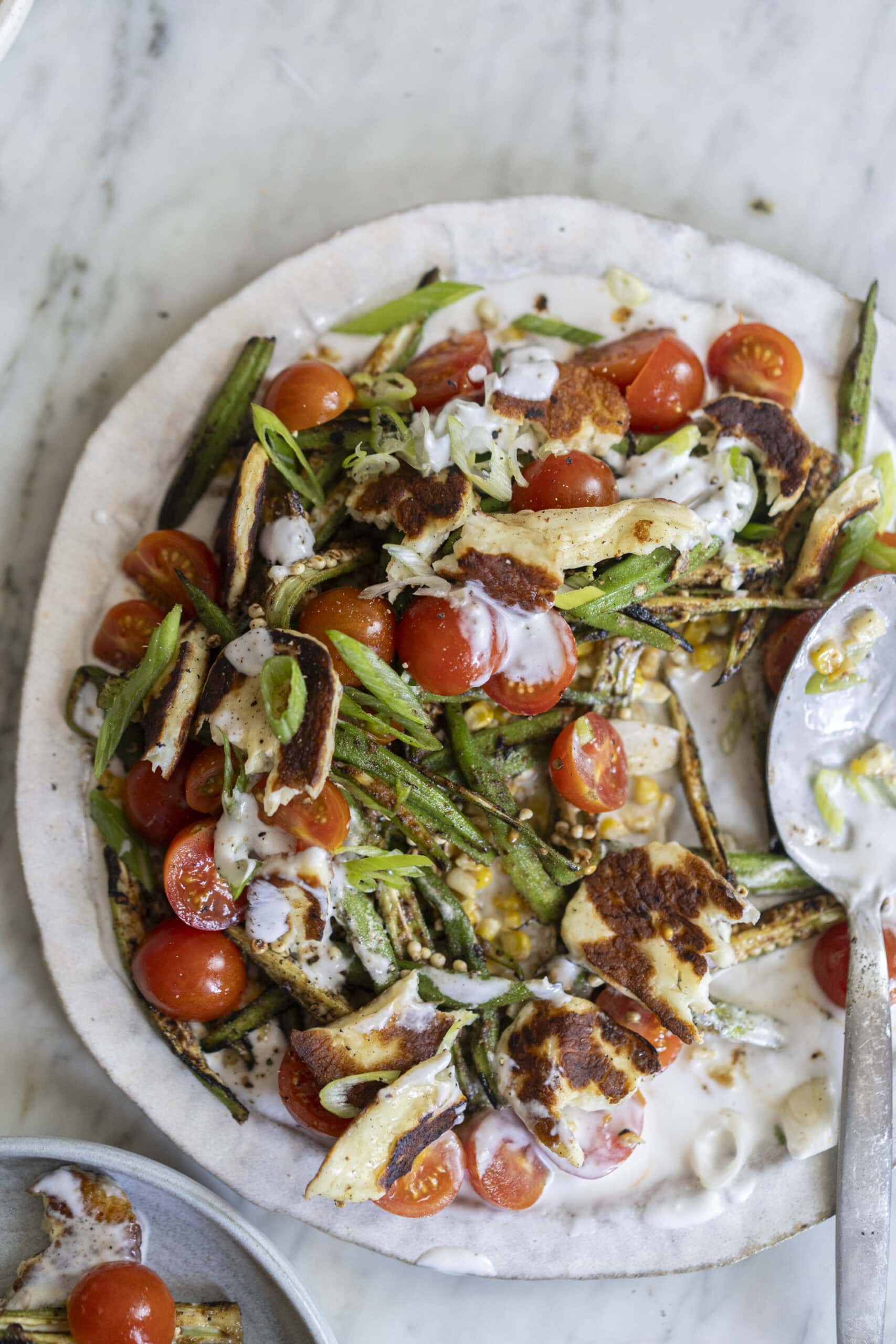 A plate of grilled halloumi, green beans, and cherry tomatoes topped with a drizzle of creamy white sauce, served on a marble surface.