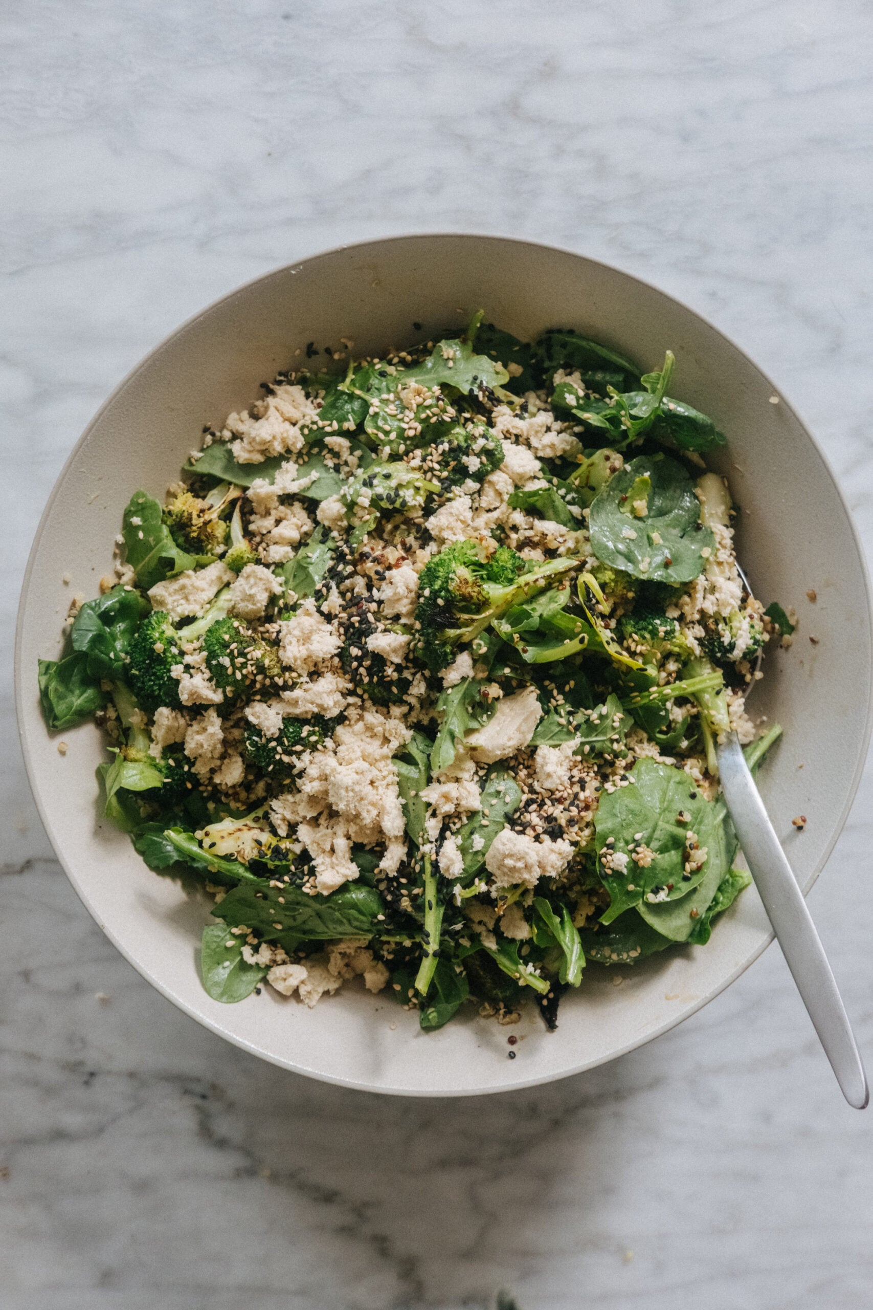 A bowl of mixed green salad with crumbled cheese, seeds, and a metal serving spoon on a marble surface.