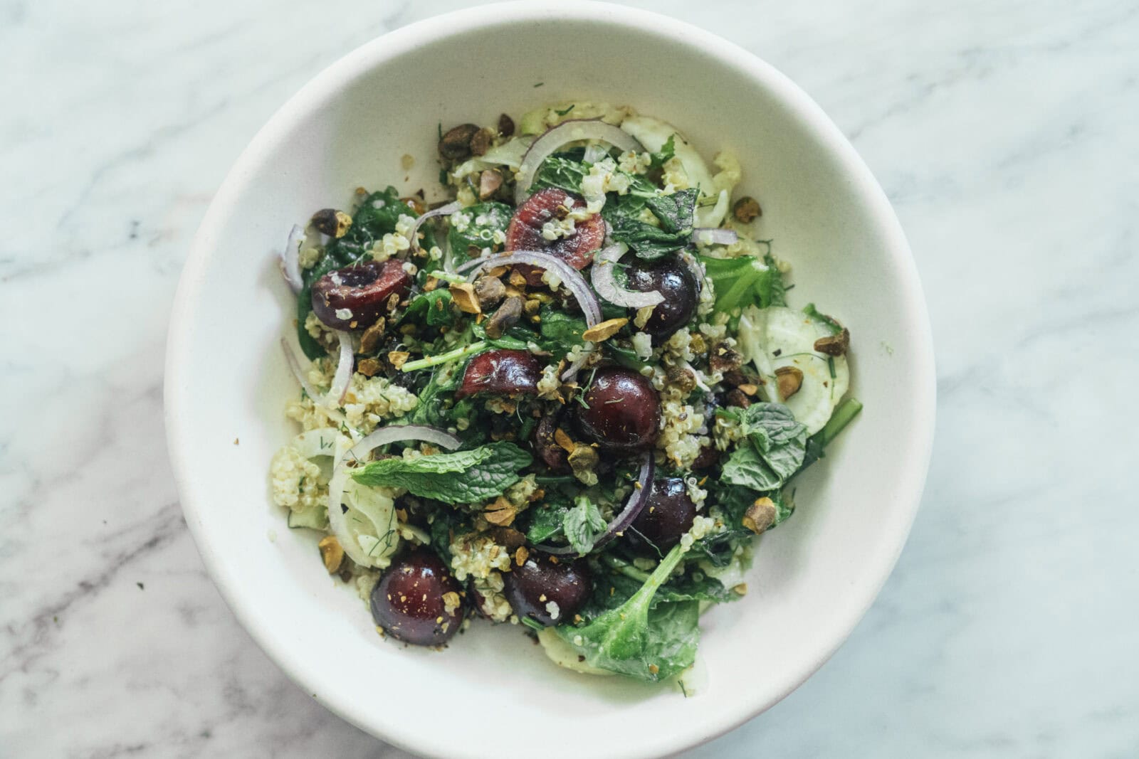 A bowl of salad with leafy greens, red onions, cherries, quinoa, herbs, and chopped nuts on a marble surface.