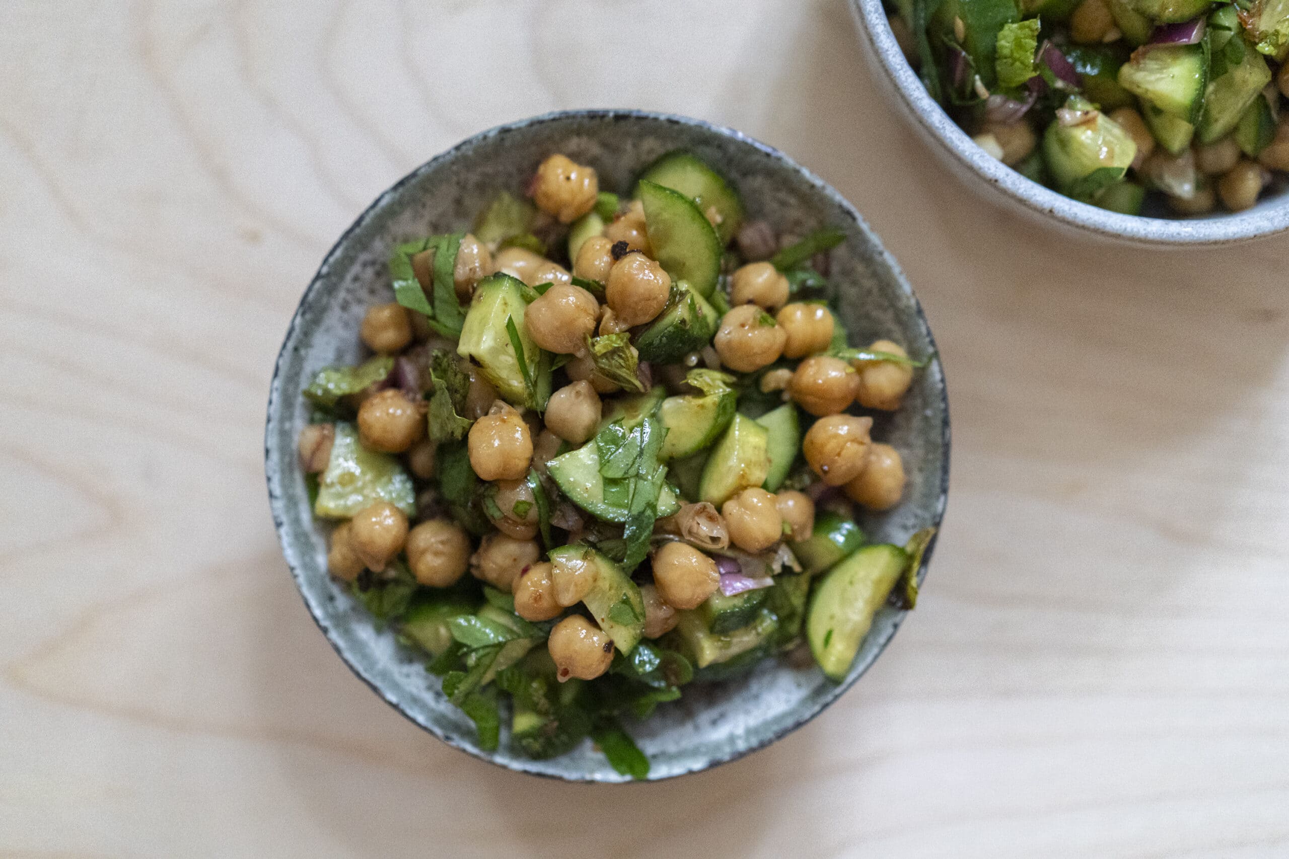 A bowl of chickpea salad with chopped cucumber, parsley, and red onion on a light wooden surface.