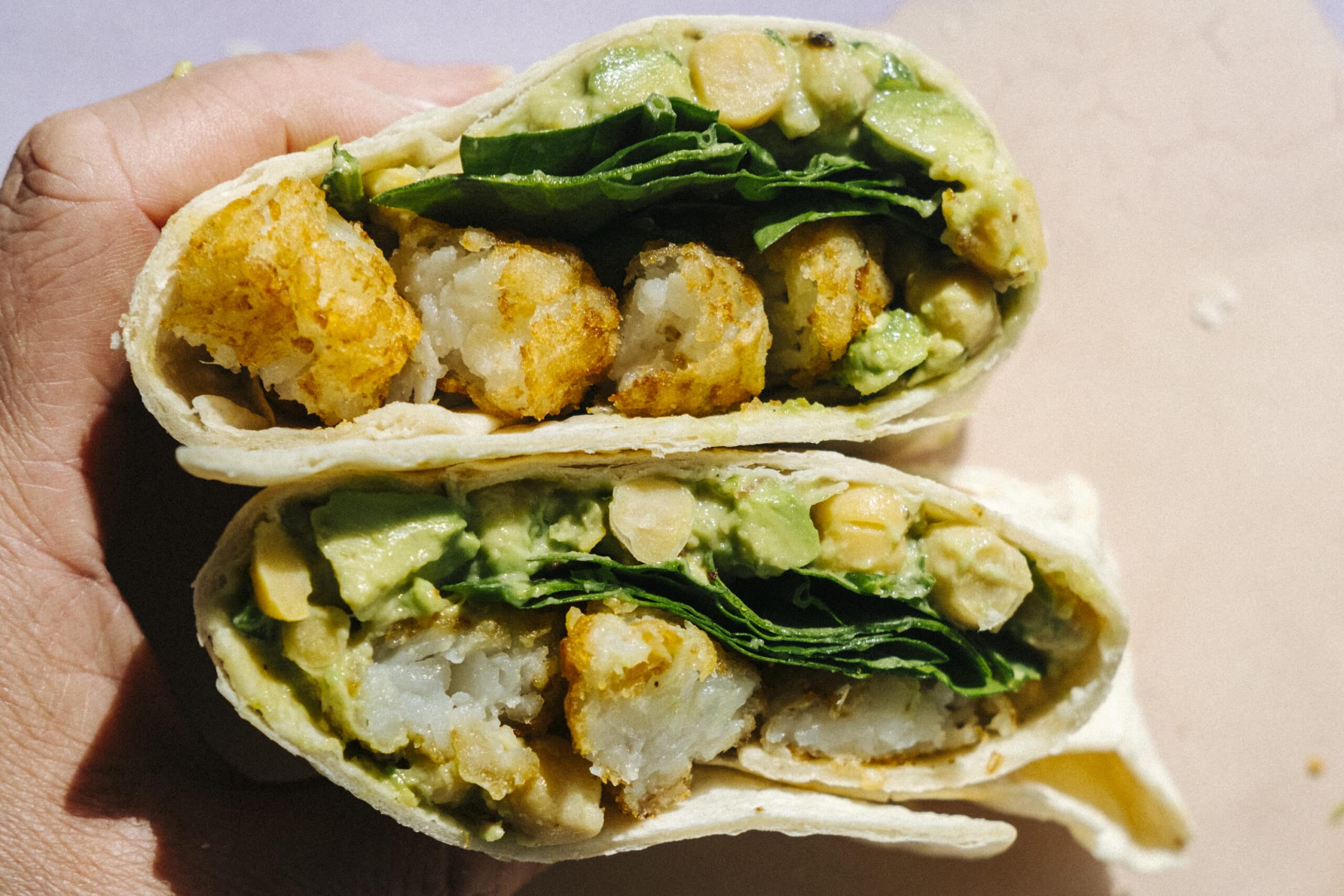 A hand holds two halves of a wrap filled with spinach, chickpeas, avocado, and crispy potato bites.