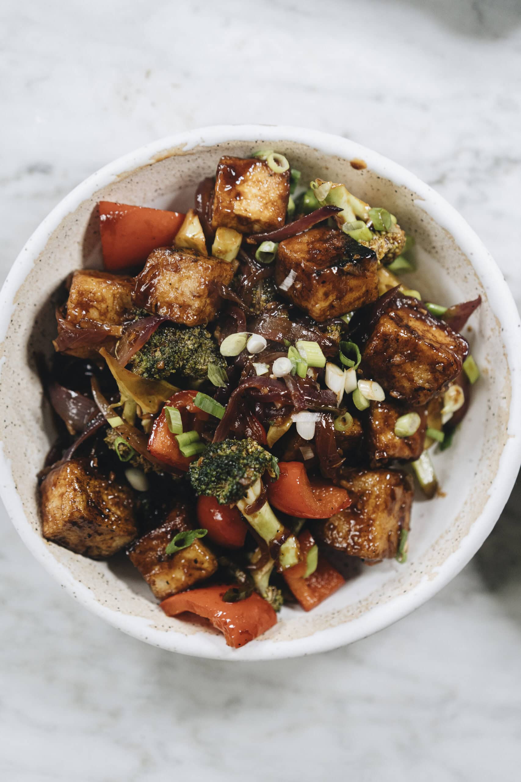 A bowl of stir-fried tofu with broccoli, red bell peppers, onions, and green onions, topped with sauce.