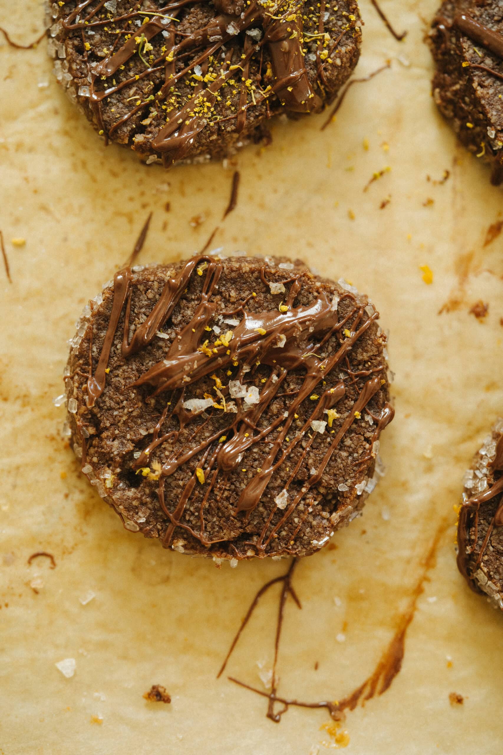 A round chocolate cookie topped with drizzled chocolate, sea salt flakes, and lemon zest on a parchment-lined surface.