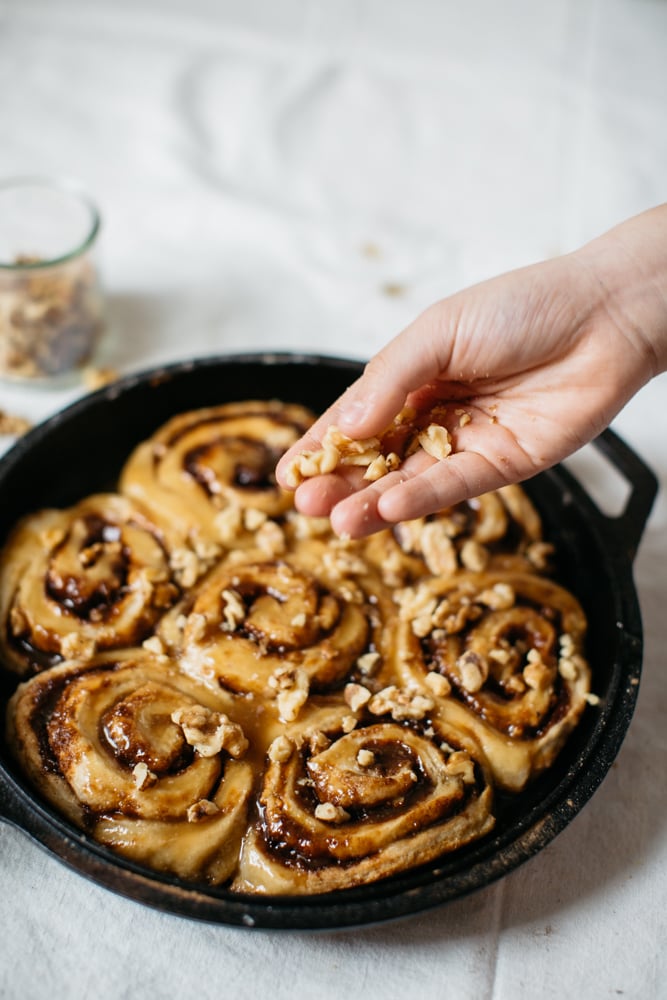 A hand sprinkles chopped walnuts over a pan of unbaked cinnamon rolls on a white surface. - Hetty Lui McKinnon