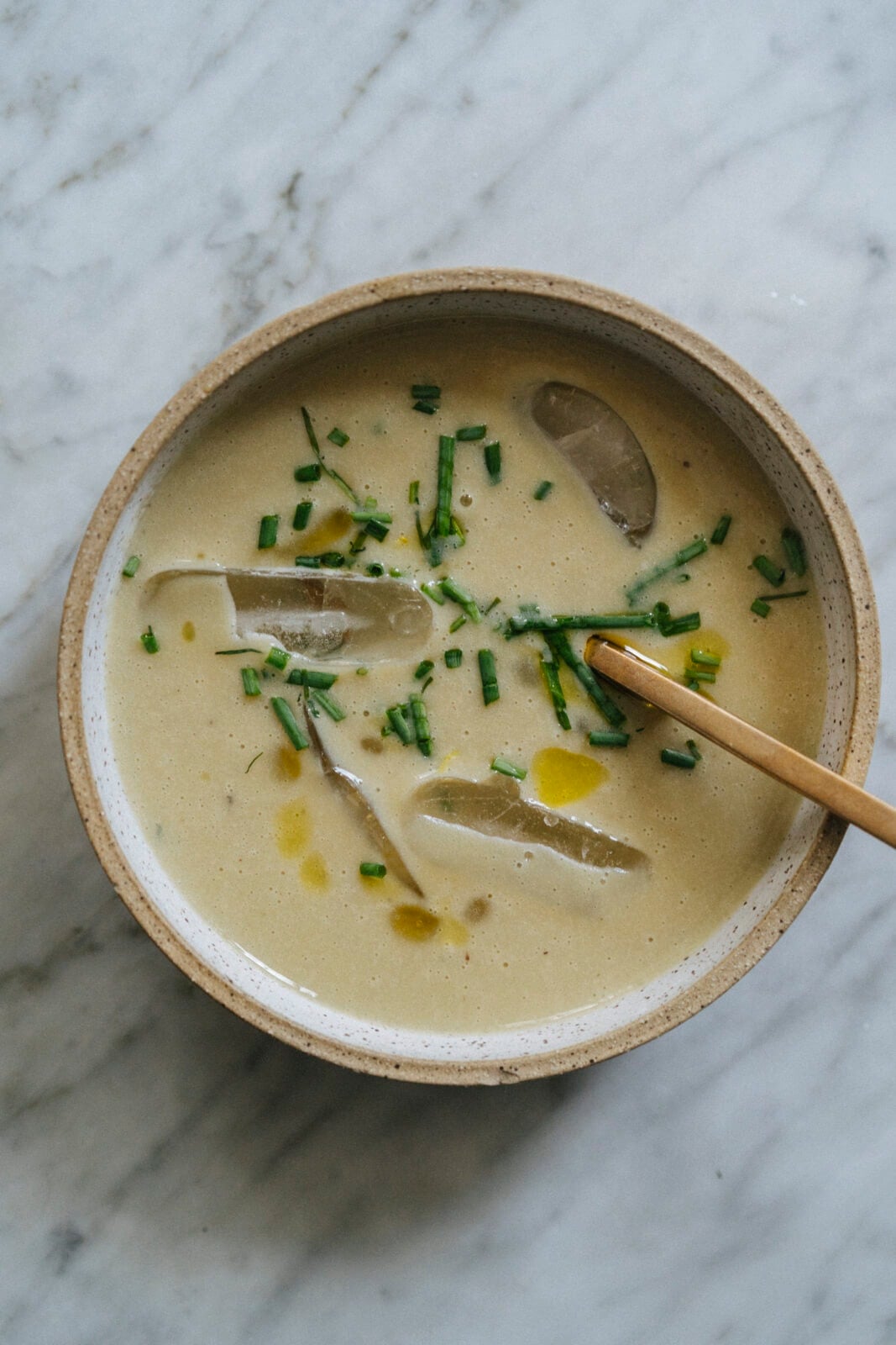 A bowl of creamy soup with ice cubes, chopped chives, and a drizzle of oil, served with a wooden spoon on a marble surface. - Hetty Lui McKinnon