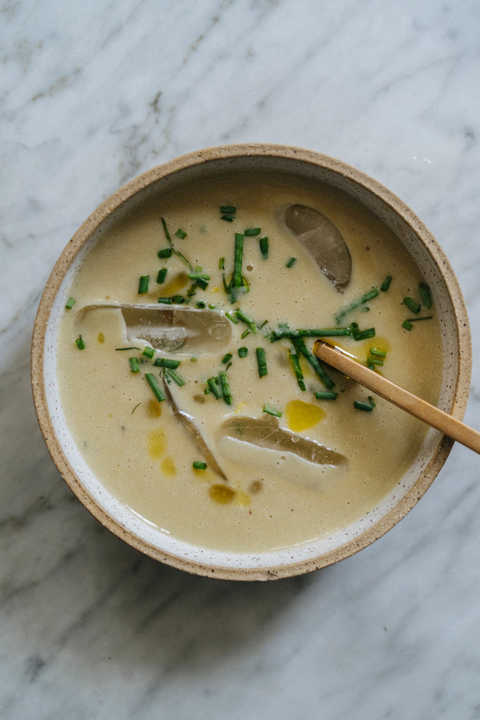 A bowl of creamy soup with ice cubes, chopped chives, and a drizzle of oil, served with a wooden spoon on a marble surface. - Hetty Lui McKinnon