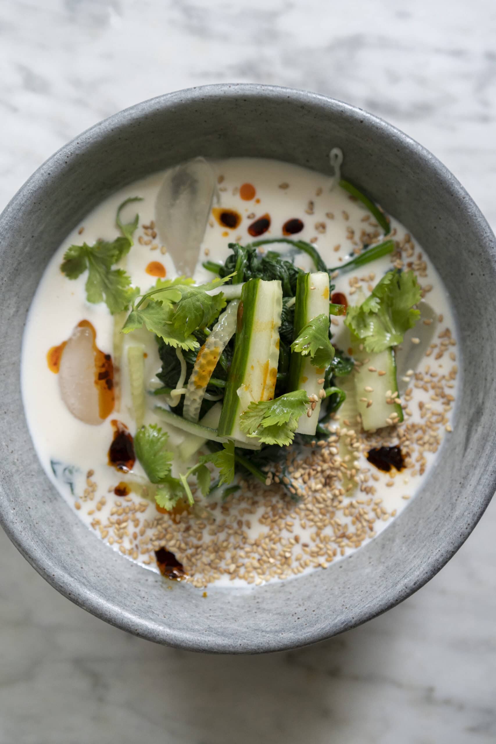 A gray bowl filled with a creamy soup, topped with sliced cucumbers, cilantro, sesame seeds, leafy greens, and a drizzle of chili oil.