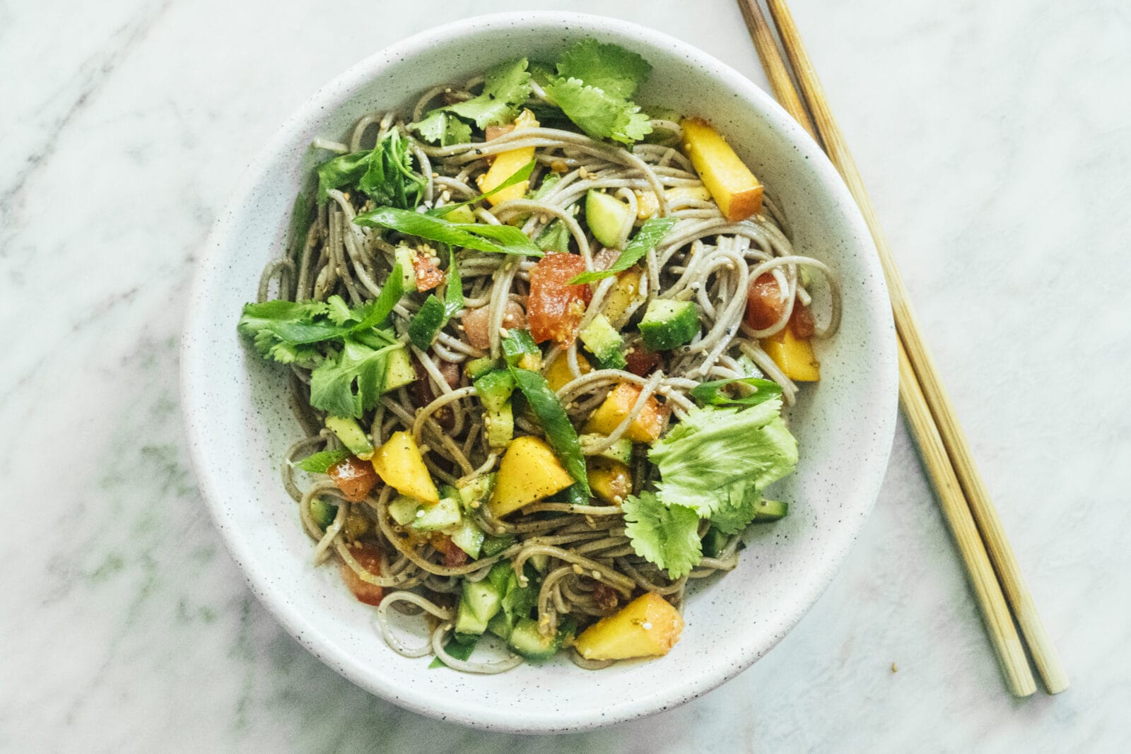 A bowl of soba noodle salad with chopped mango, cucumber, tomato, and cilantro on a marble surface; wooden chopsticks are beside the bowl. - Hetty Lui McKinnon