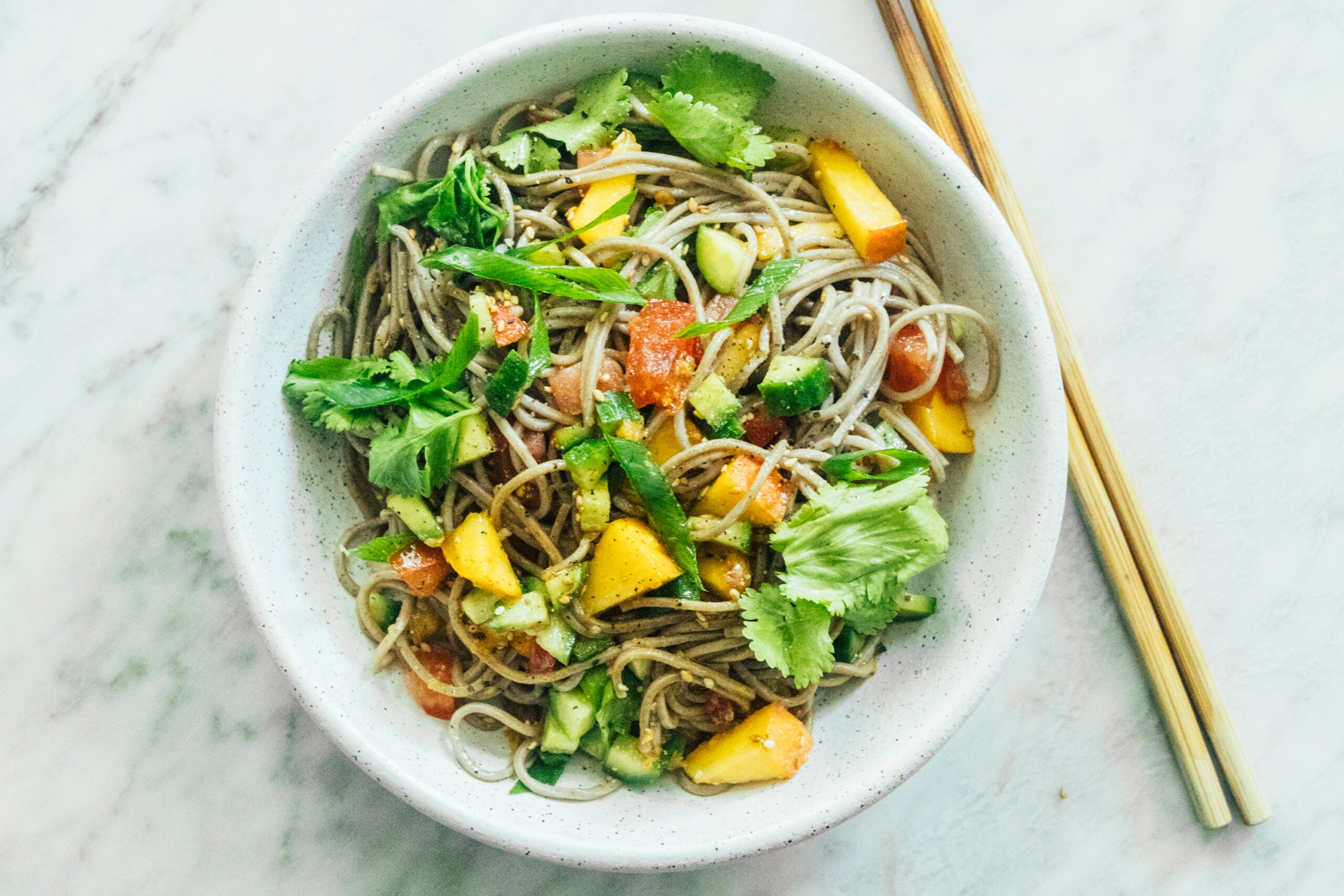 A bowl of soba noodle salad with chopped mango, cucumber, tomato, and cilantro on a marble surface; wooden chopsticks are beside the bowl. - Hetty Lui McKinnon