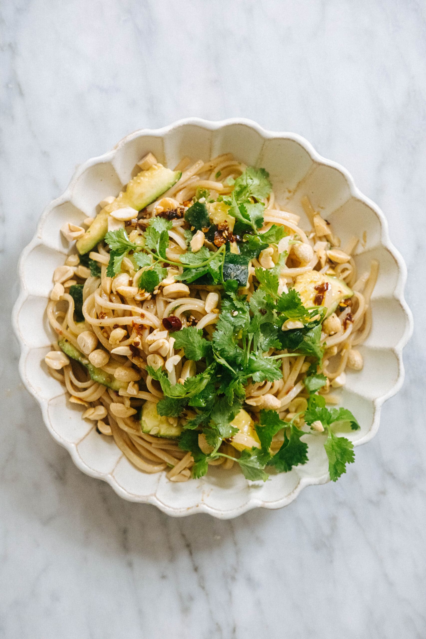 A bowl of noodles topped with chopped peanuts, cilantro, zucchini, and a light sauce, served in a scalloped white dish on a marble surface.