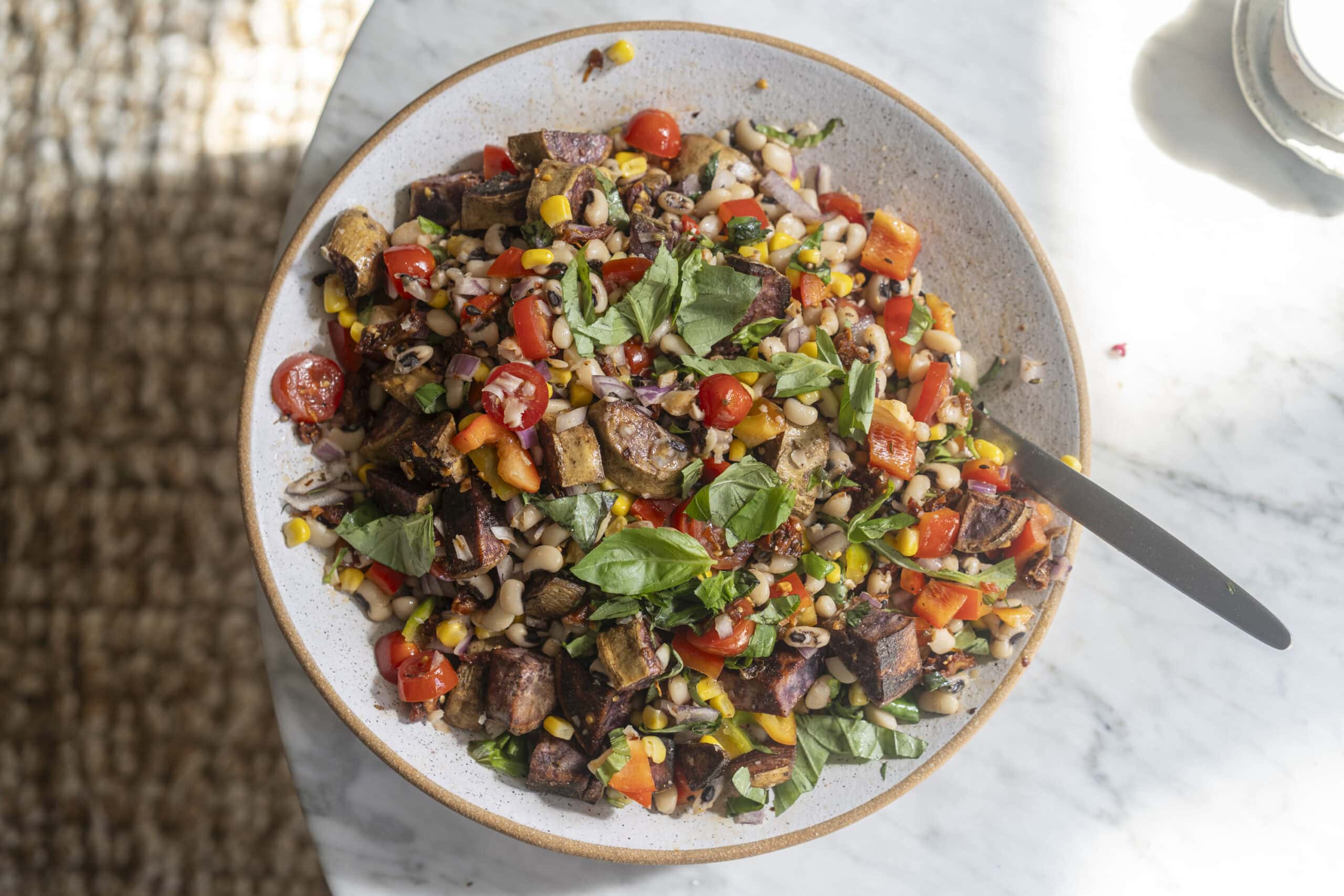 A bowl of mixed salad with black-eyed peas, cherry tomatoes, corn, red bell peppers, sweet potatoes, and fresh greens on a marble surface with a spoon.