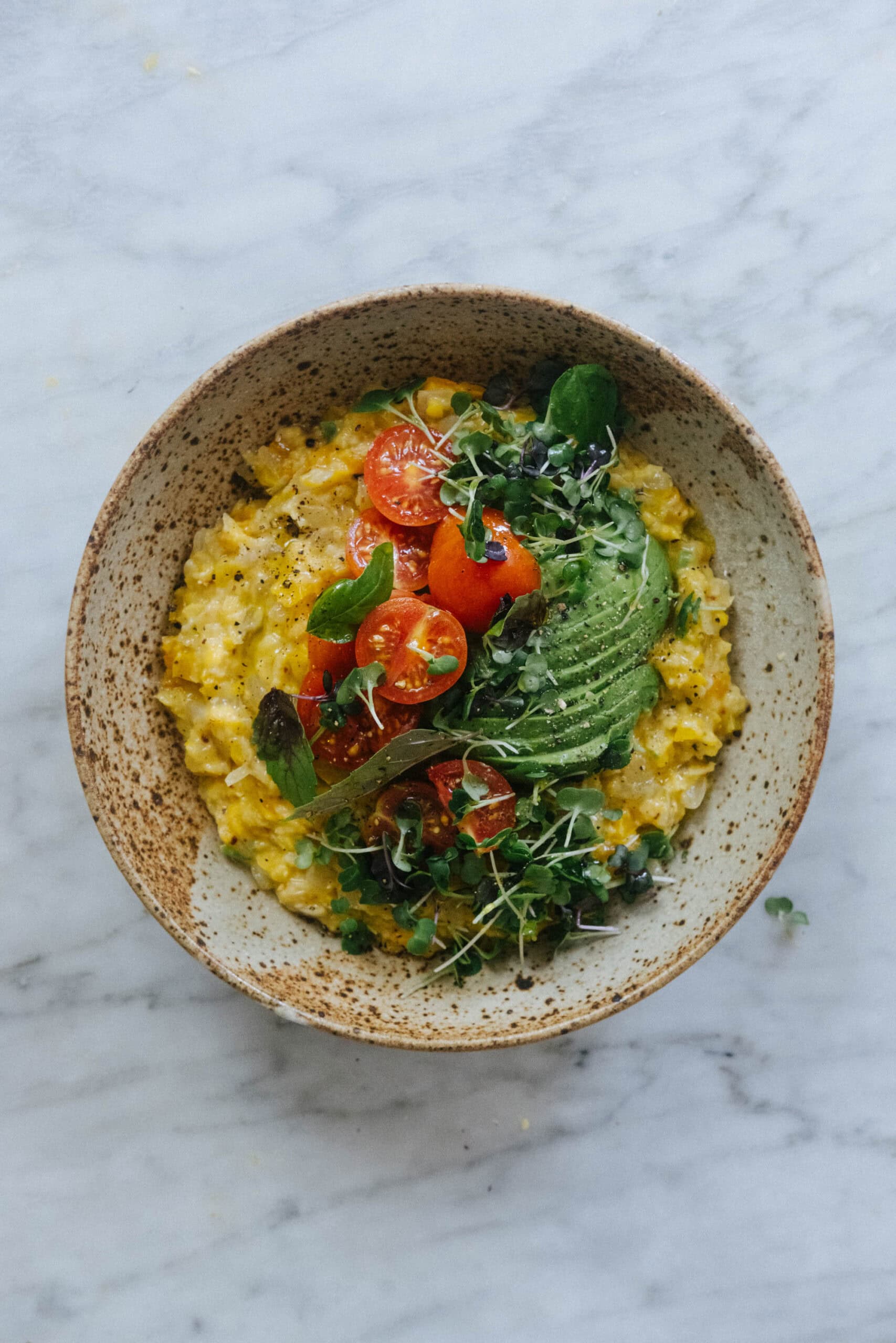 A bowl of creamy risotto topped with sliced avocado, halved cherry tomatoes, fresh basil leaves, and microgreens on a marble surface.