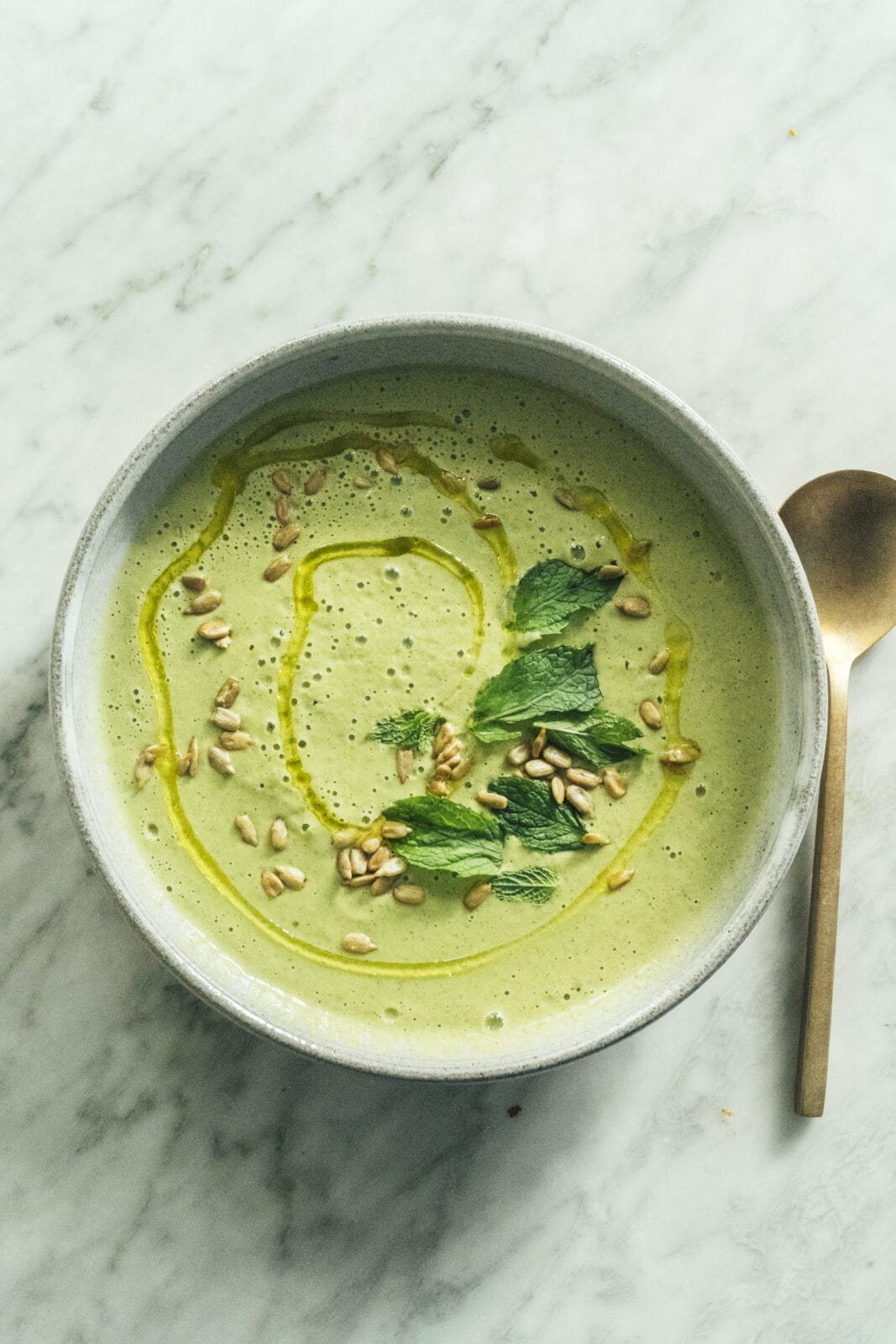 A bowl of green soup garnished with mint leaves, sunflower seeds, and a drizzle of oil, placed on a marble surface next to a spoon. - Hetty Lui McKinnon