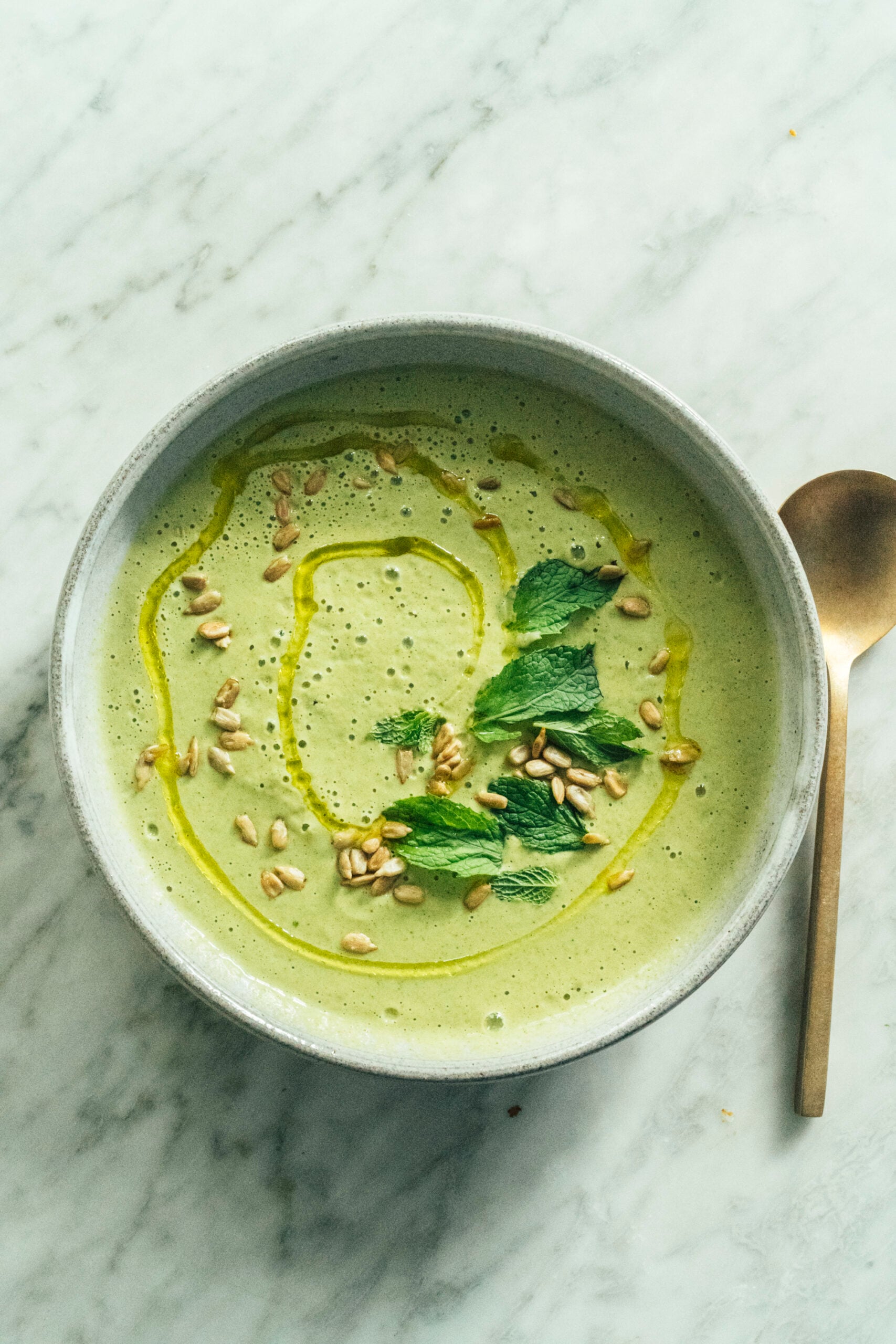 A bowl of green soup garnished with mint leaves, sunflower seeds, and a drizzle of oil, placed on a marble surface next to a spoon. - Hetty Lui McKinnon