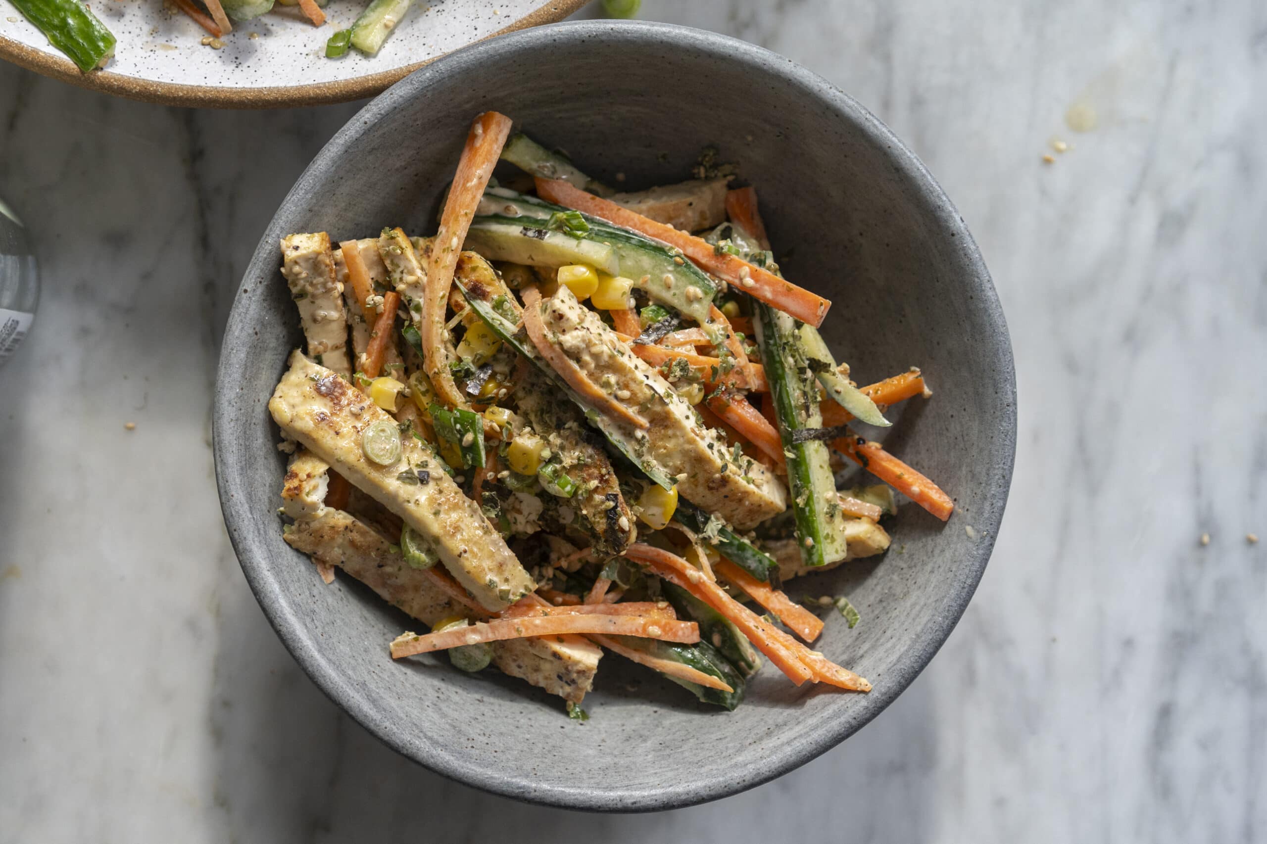 A bowl of salad with sliced tofu, carrots, cucumbers, corn, and herbs on a marble surface.