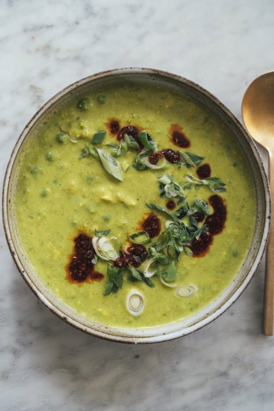 A bowl of green soup garnished with chopped green onions and chili oil, placed on a marble surface with a gold spoon beside it.