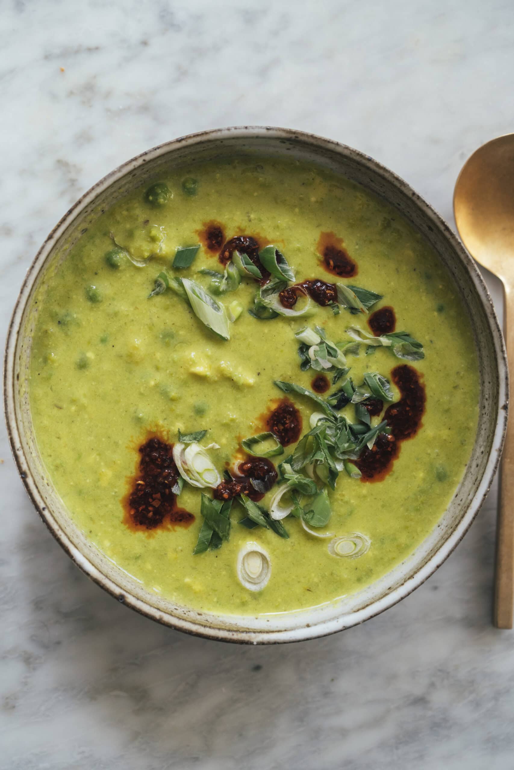 A bowl of green soup garnished with chopped green onions and chili oil, placed on a marble surface with a gold spoon beside it.