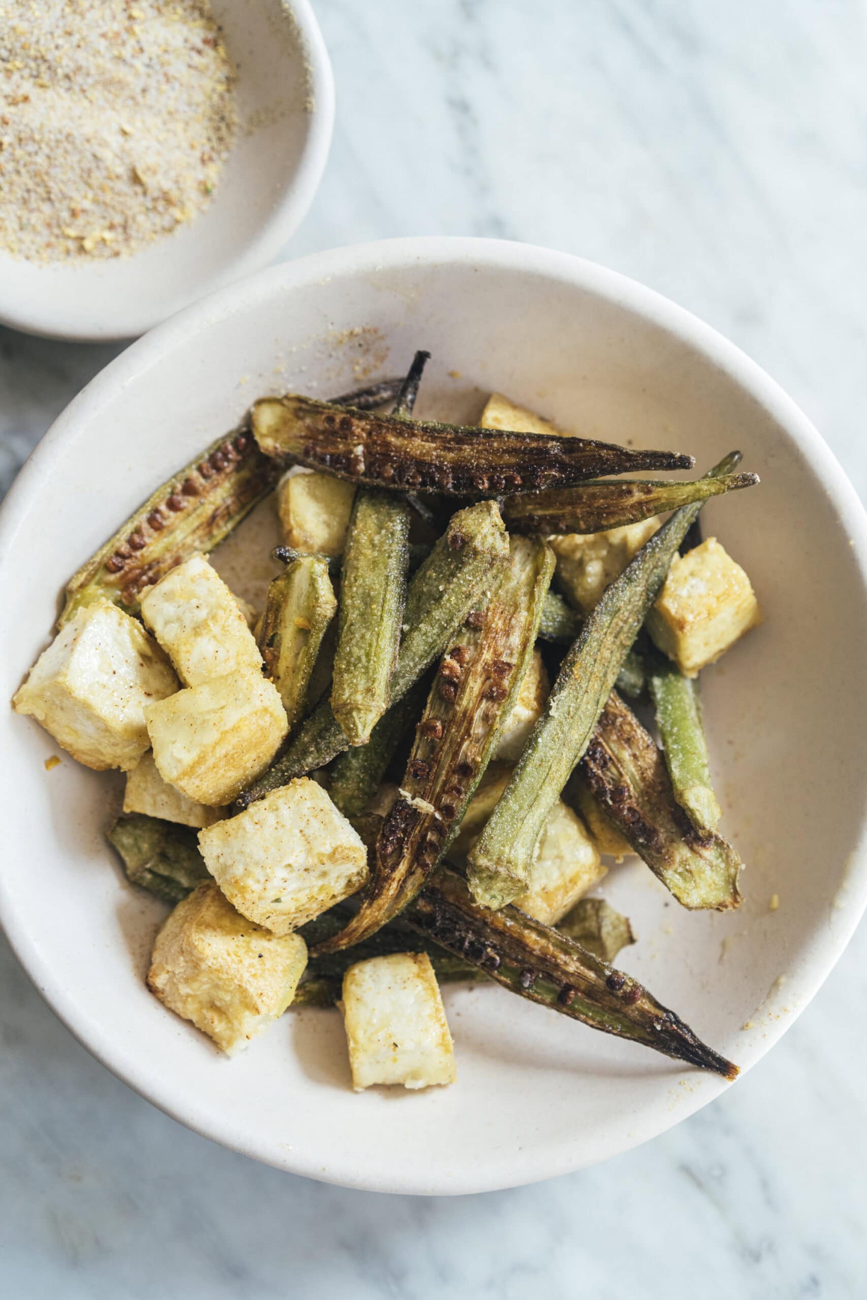 A white bowl containing roasted okra and tofu cubes, with a small dish of seasoning visible in the background on a marble surface.